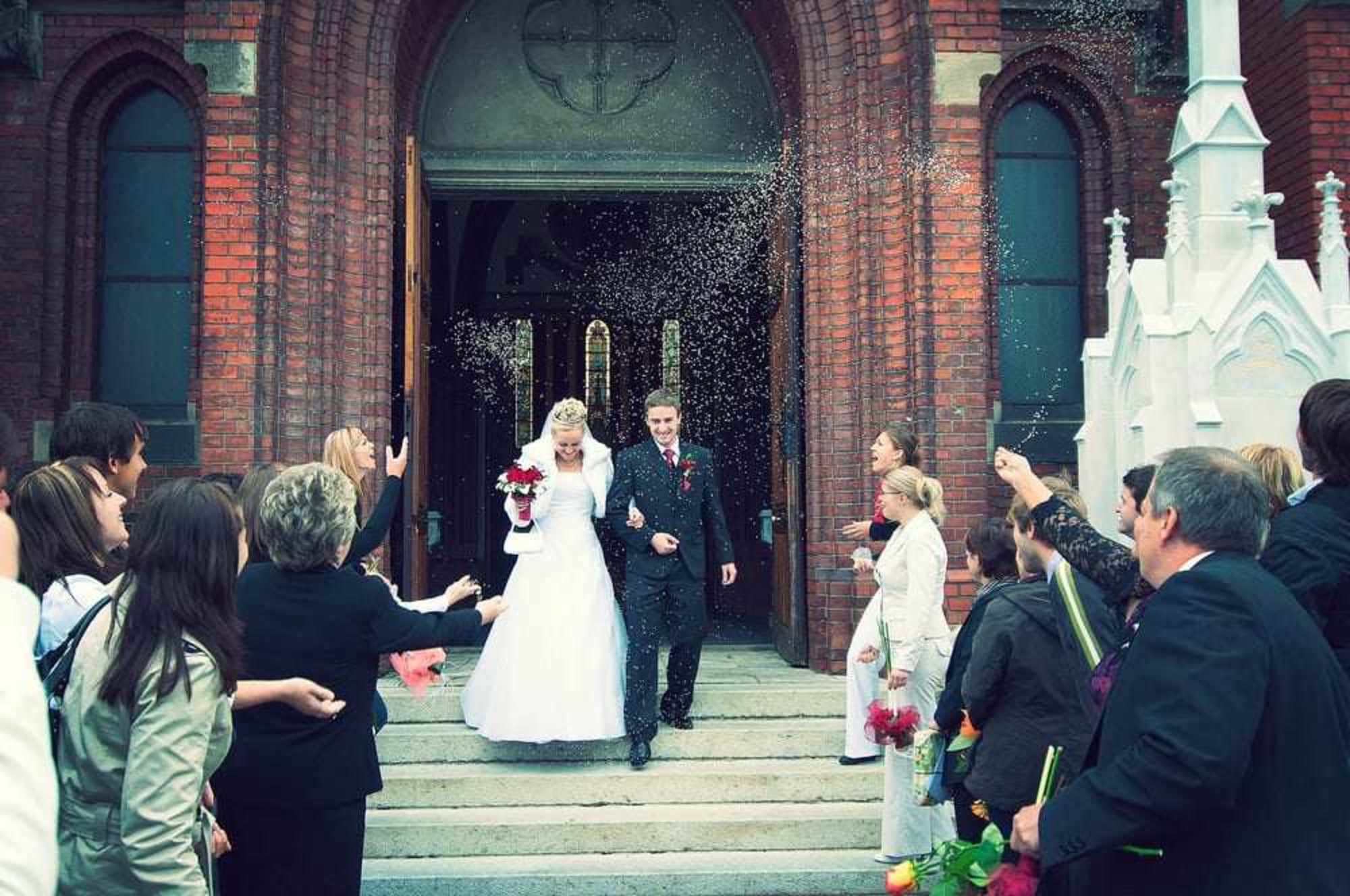 Bride and groom walk out of a brick church as guests throw confetti and celebrate around them.