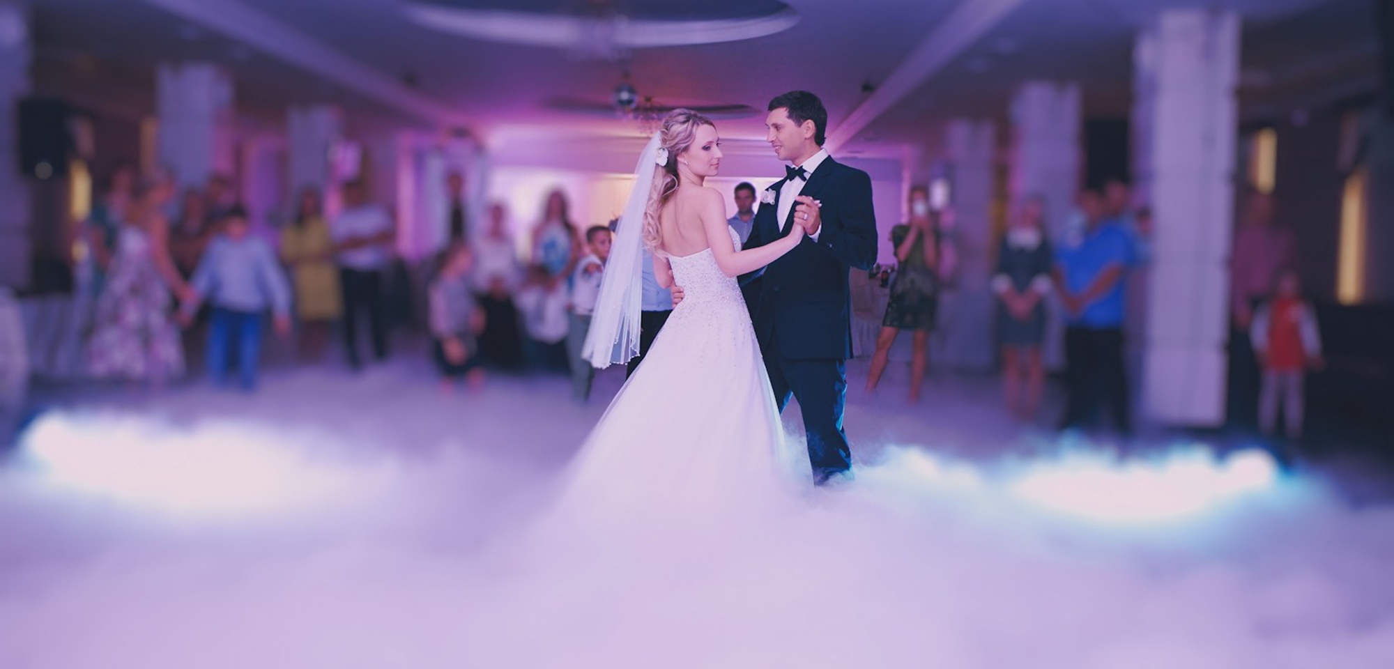 Bride and groom share their first dance on a fog-covered dance floor surrounded by guests.