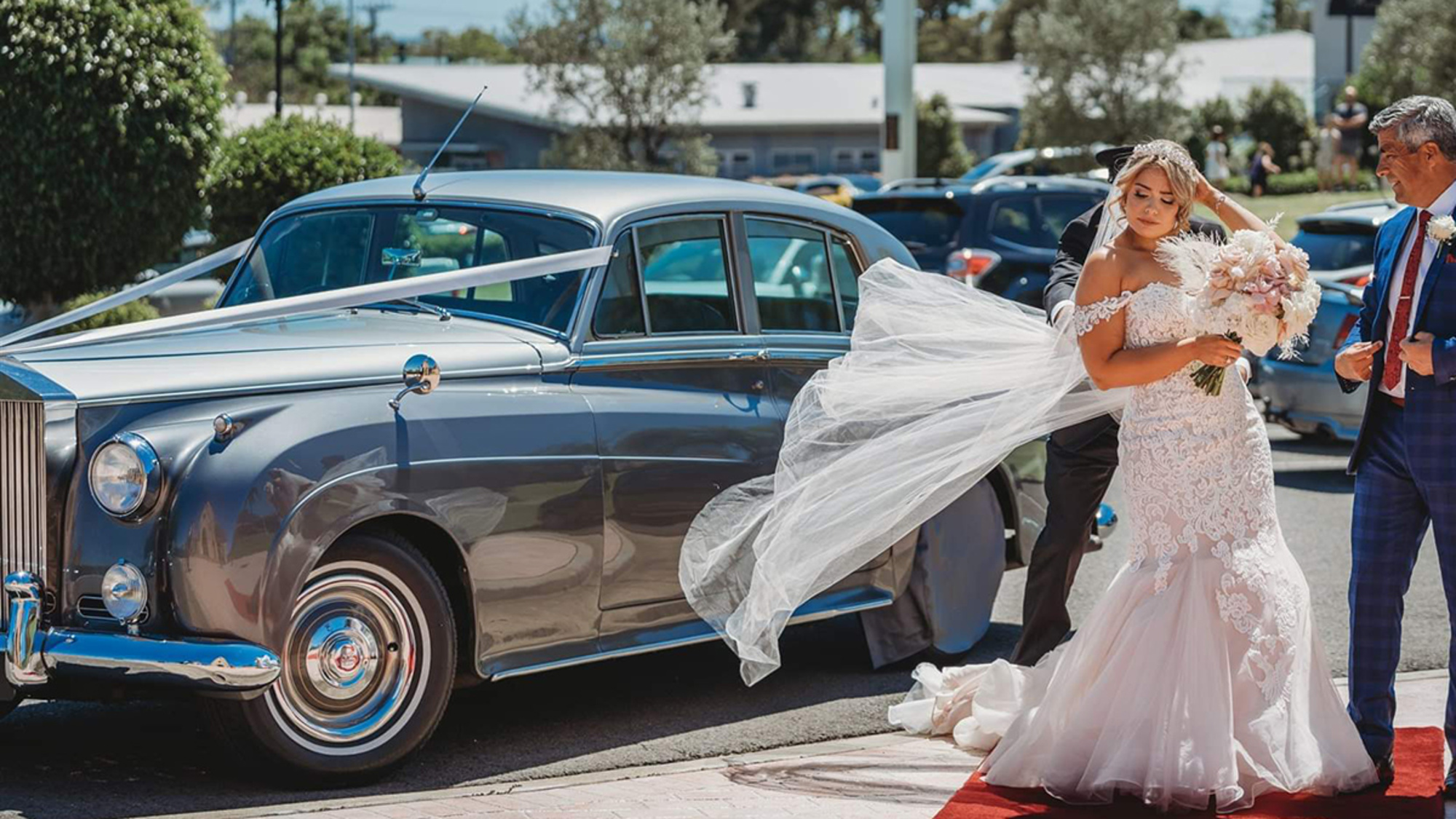 Bride in lace gown steps from a classic silver wedding car with flowing veil and bouquet on a red carpet.