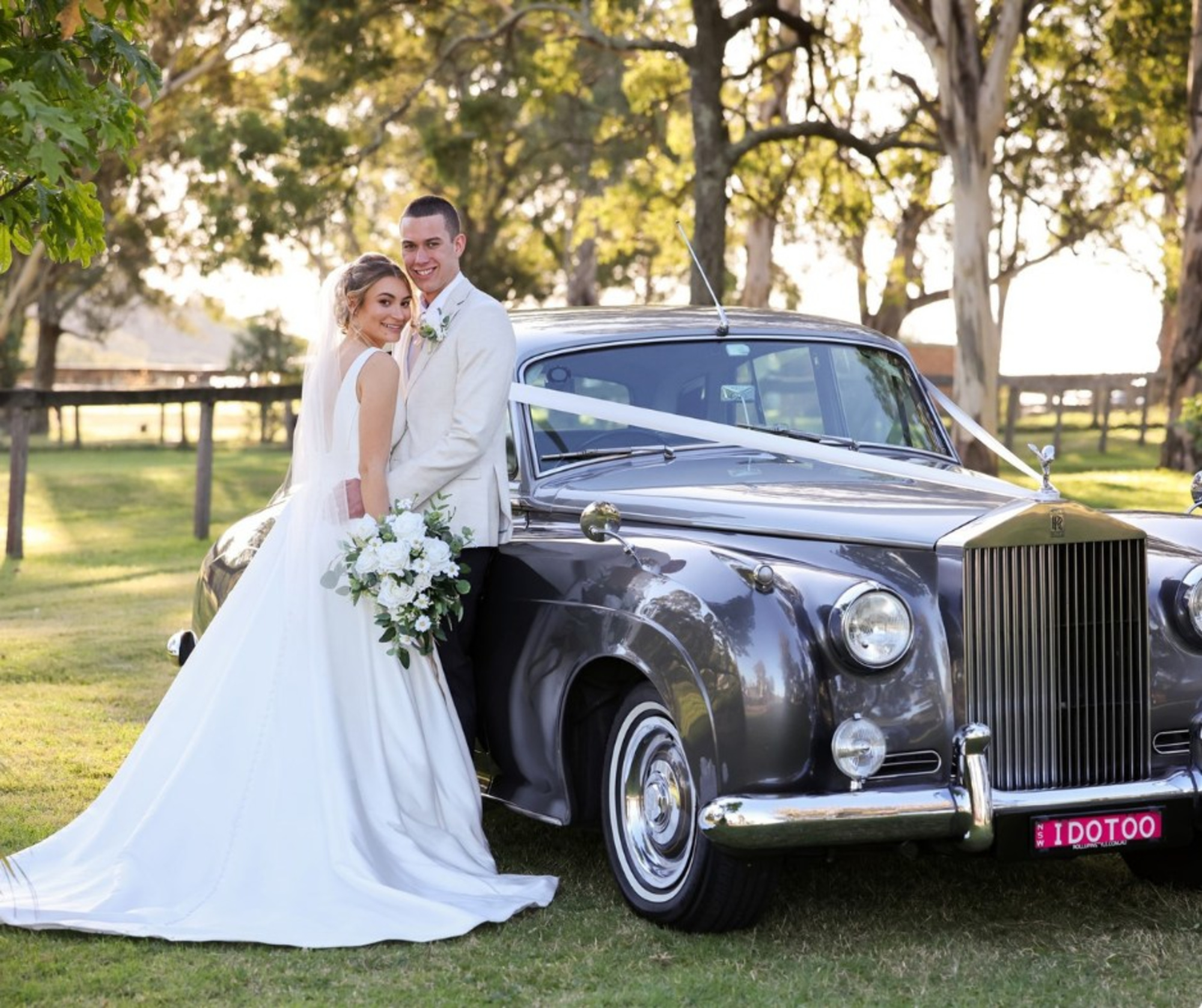 Bride and groom pose beside a classic Rolls Royce wedding car in a sunny garden setting.