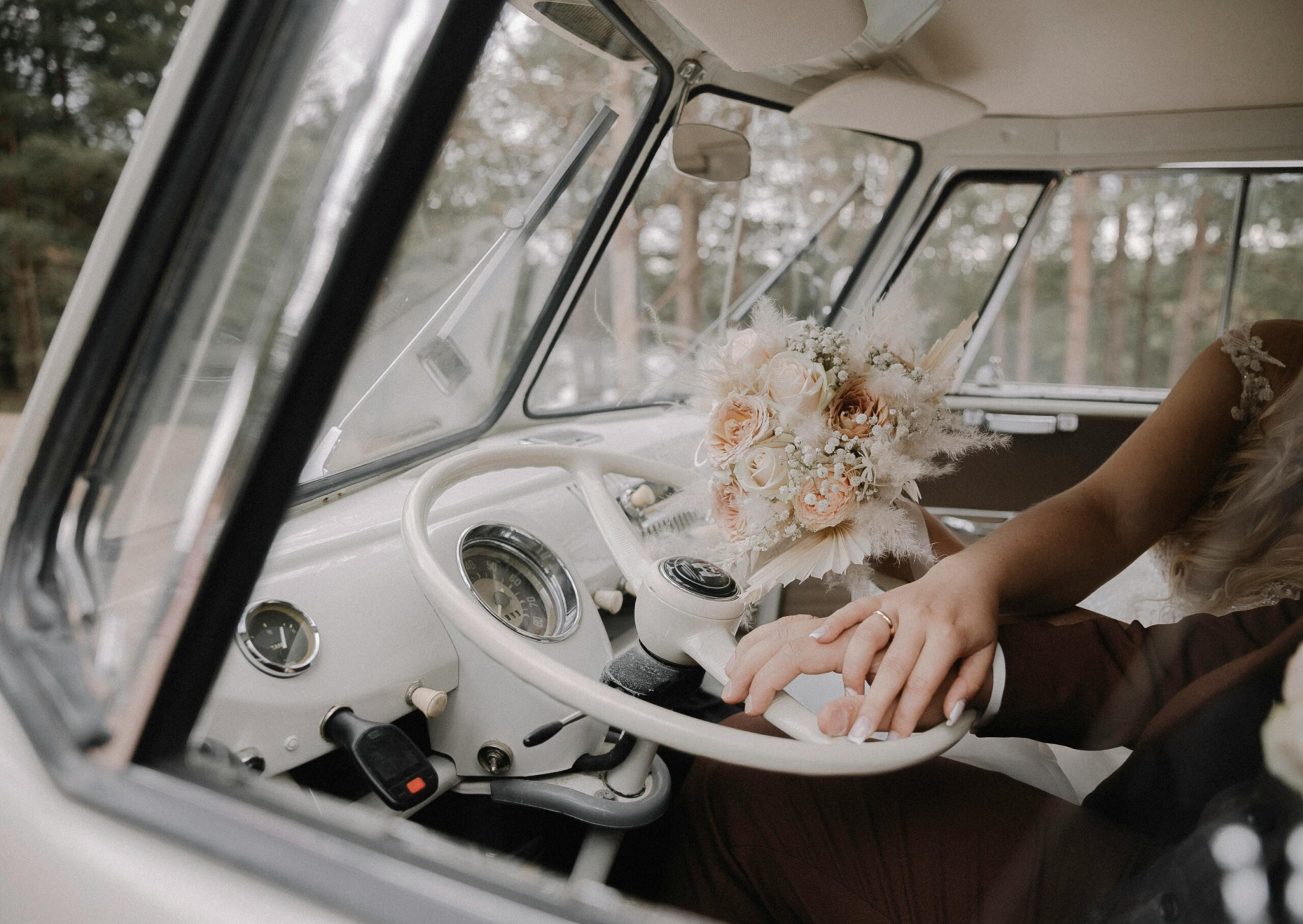 Close-up of a couple holding hands on a vintage car steering wheel with a soft blush wedding bouquet on the dashboard.