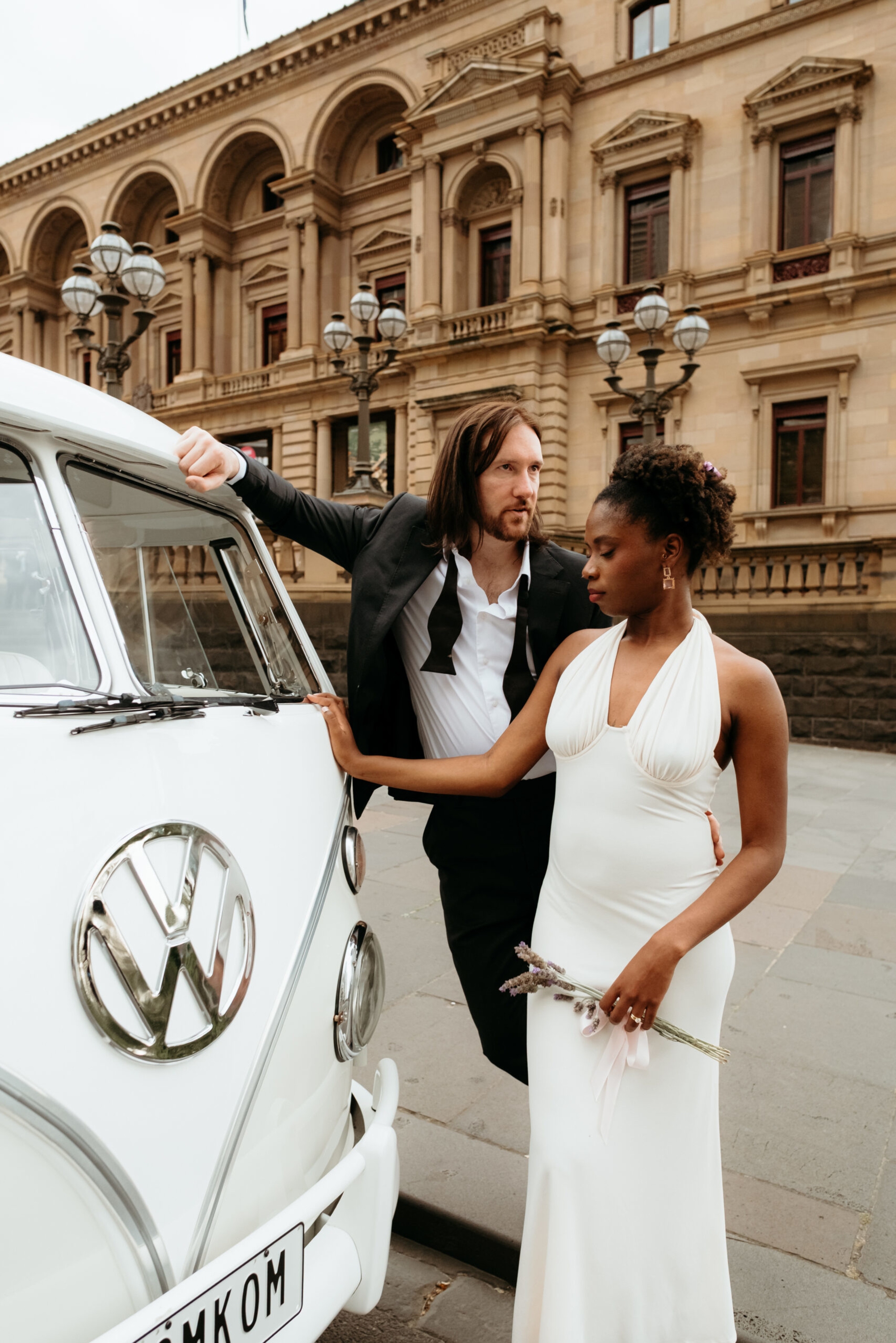 Stylish wedding couple poses beside a vintage white van in front of a grand city building.