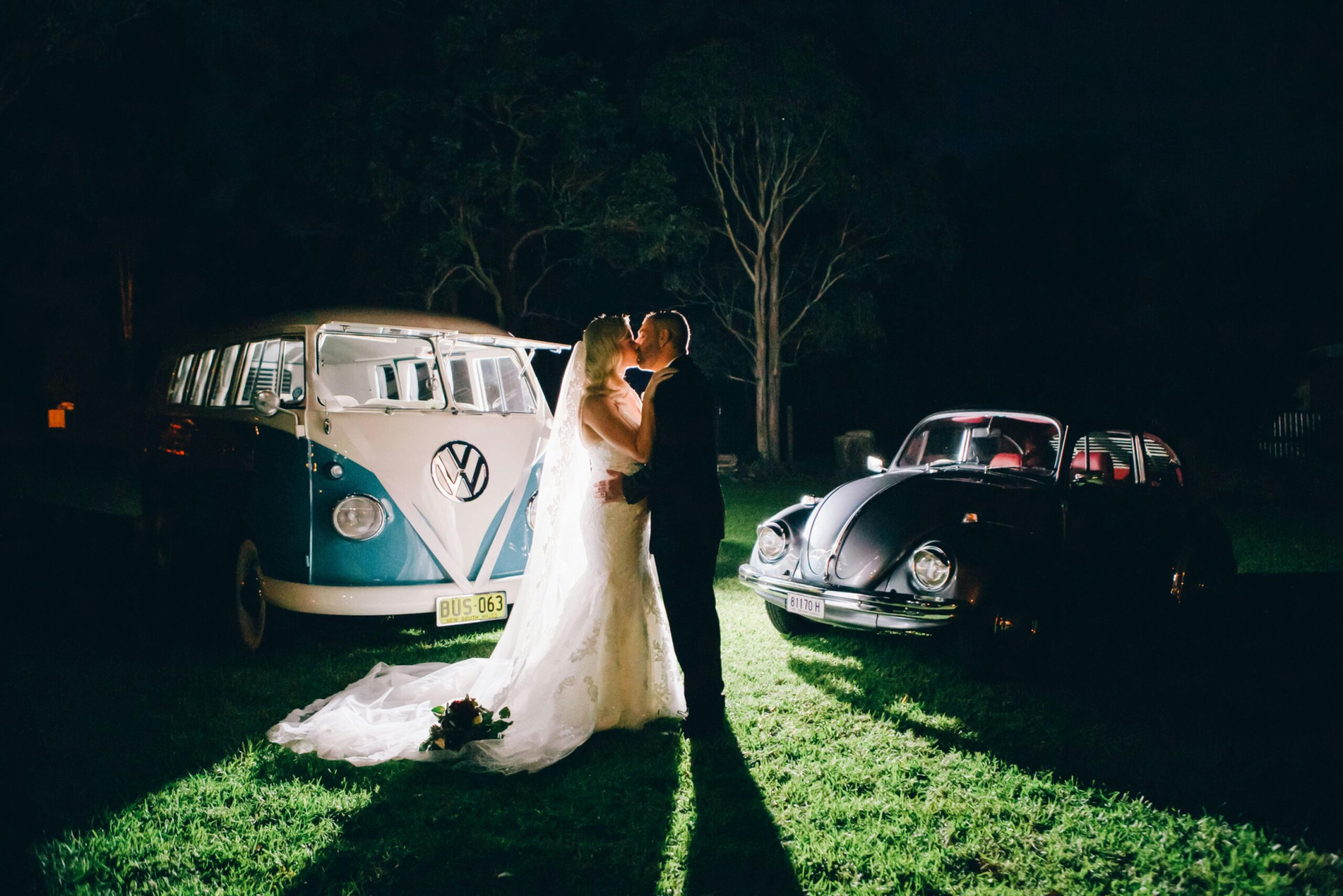 Bride and groom share a kiss at night between two vintage cars on a grassy outdoor wedding setting.