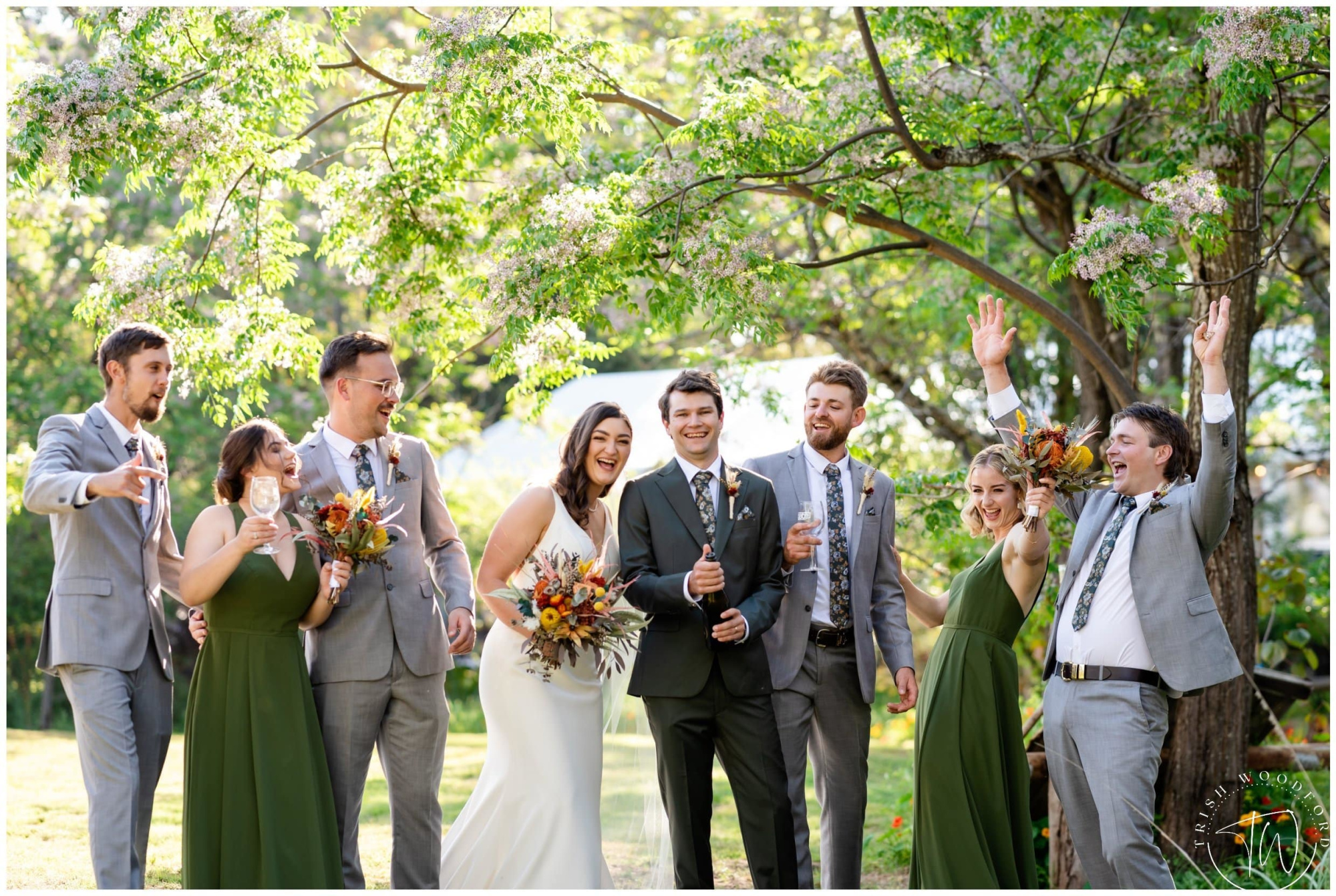 Joyful wedding party celebrating outdoors under leafy green trees with the bride and groom in the center.