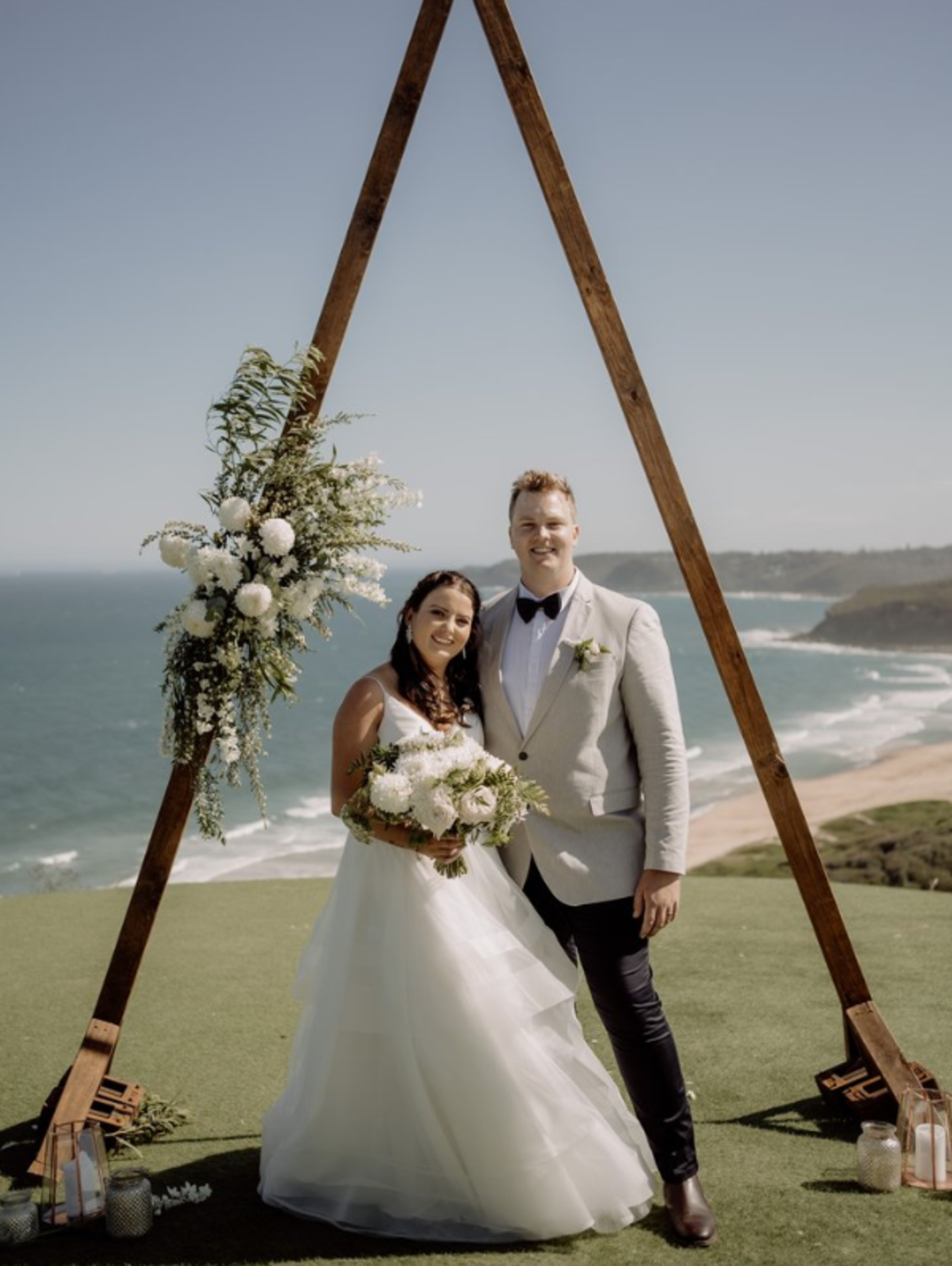 Bride and groom standing under a triangular floral arch overlooking the ocean at an outdoor coastal wedding ceremony.