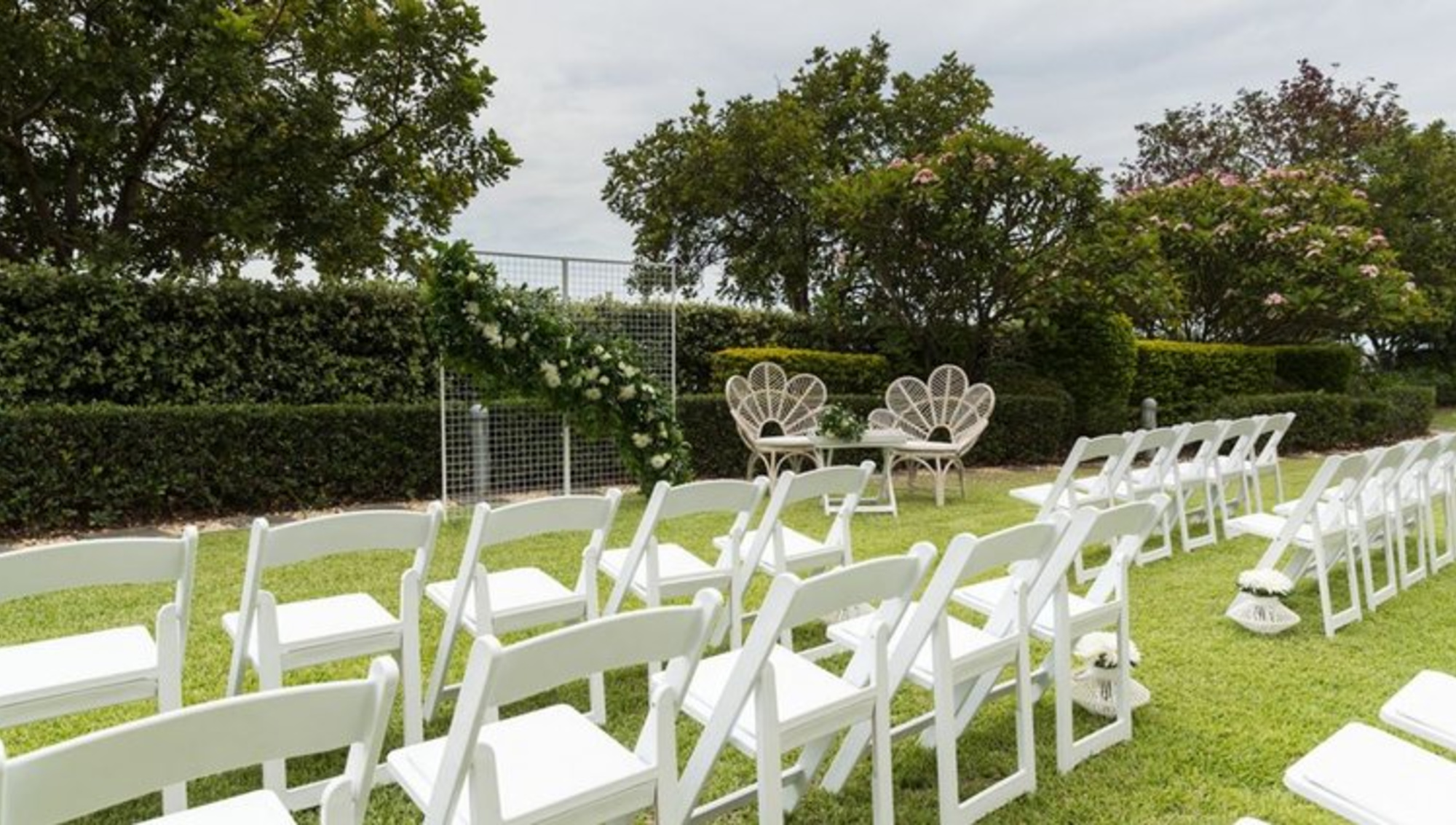 Outdoor garden wedding ceremony setup with white chairs facing a greenery backdrop and two decorative rattan chairs.