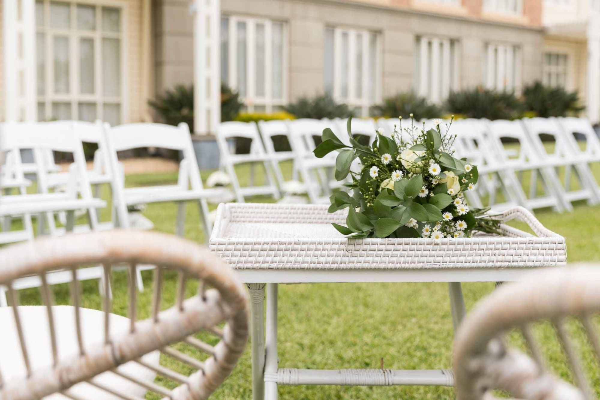 Outdoor wedding ceremony setup with white chairs and a floral arrangement on a wicker table.