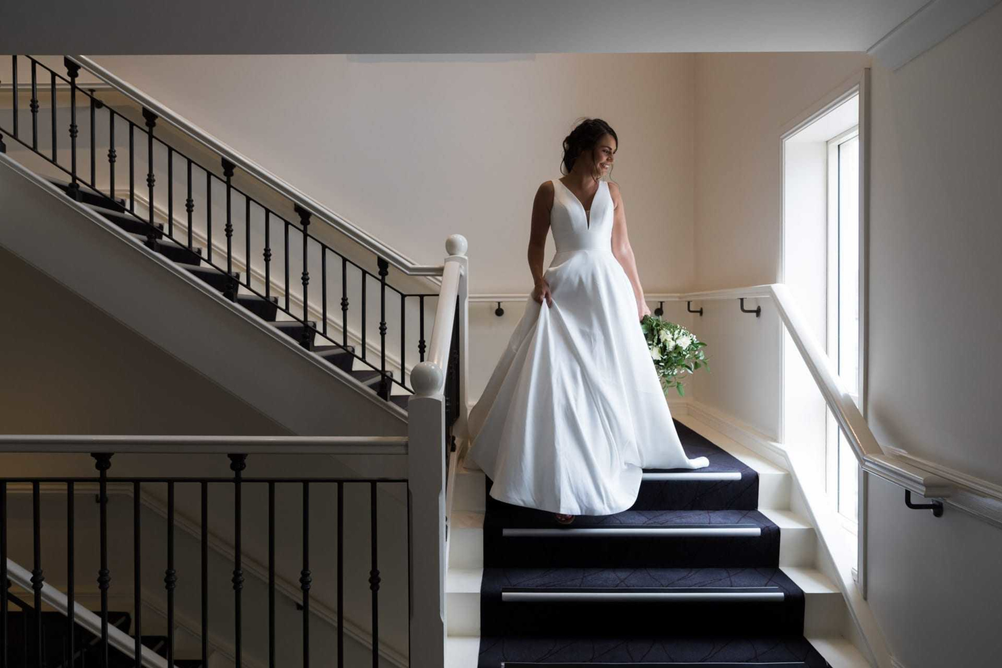 Bride in a flowing white gown descends a modern staircase holding a lush green and white bouquet.
