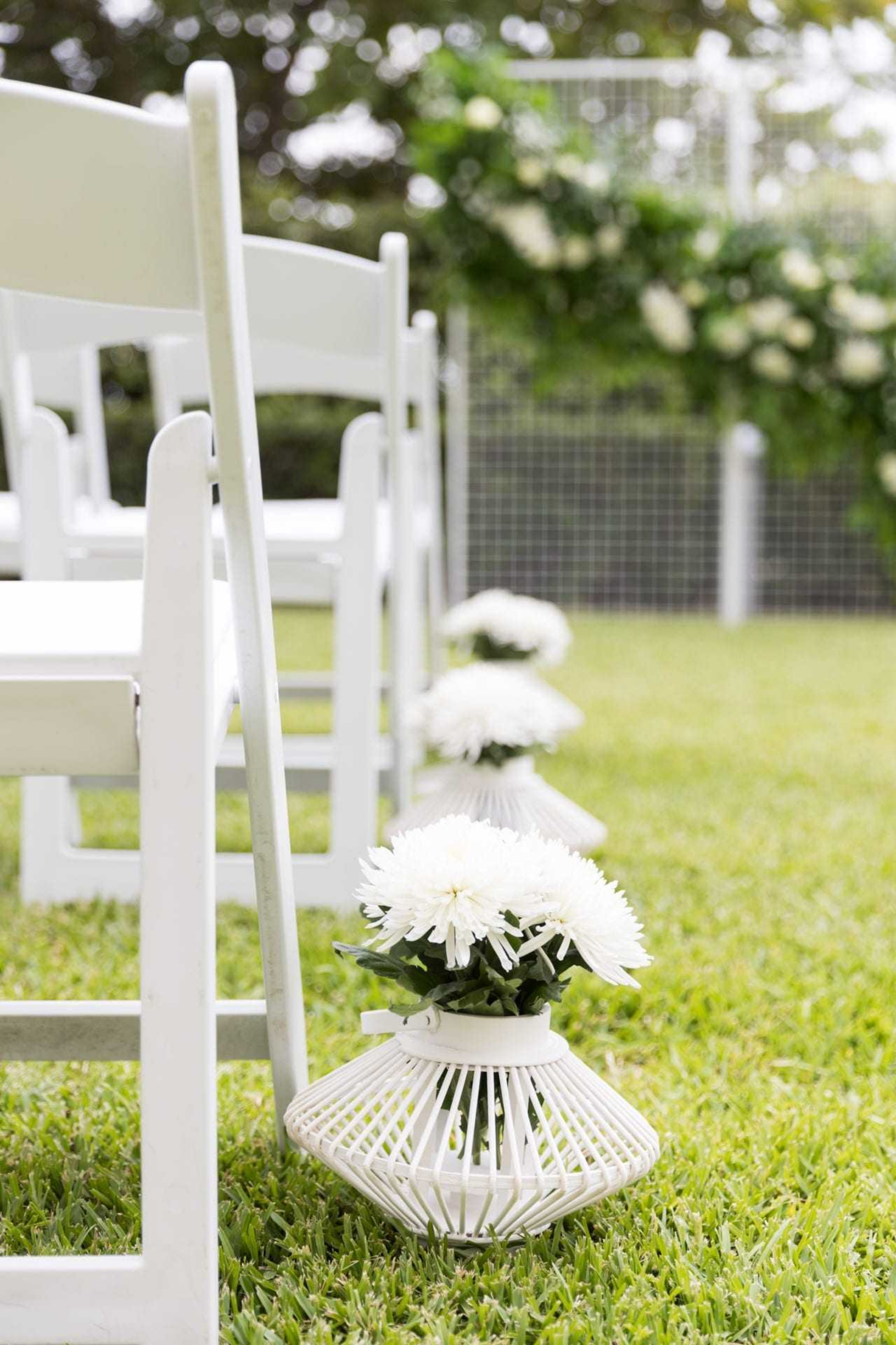 Outdoor wedding ceremony aisle with white chairs and white flower arrangements in lantern vases on green grass.