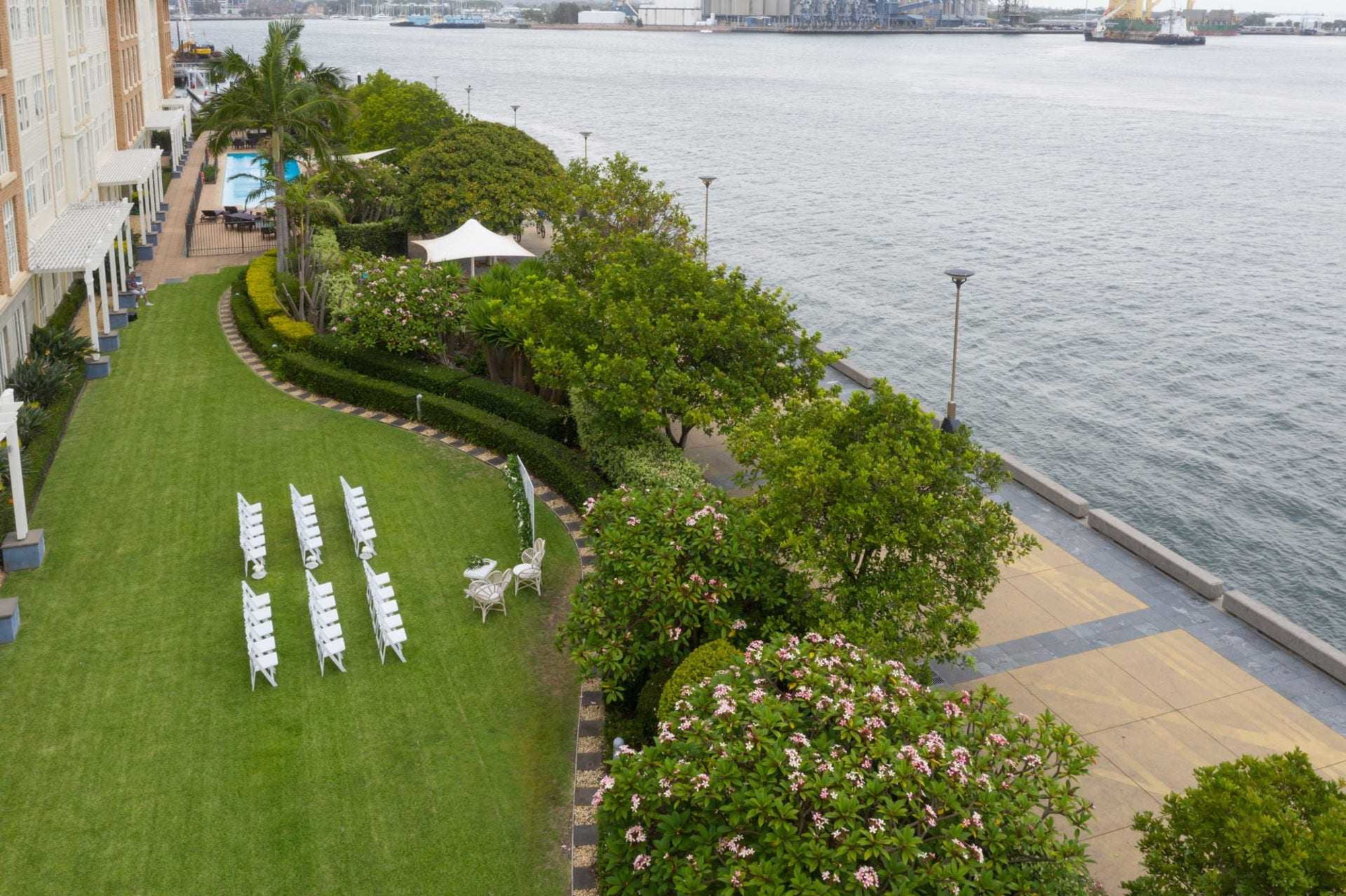 Waterfront lawn wedding ceremony setup with white chairs and greenery beside a riverside promenade.