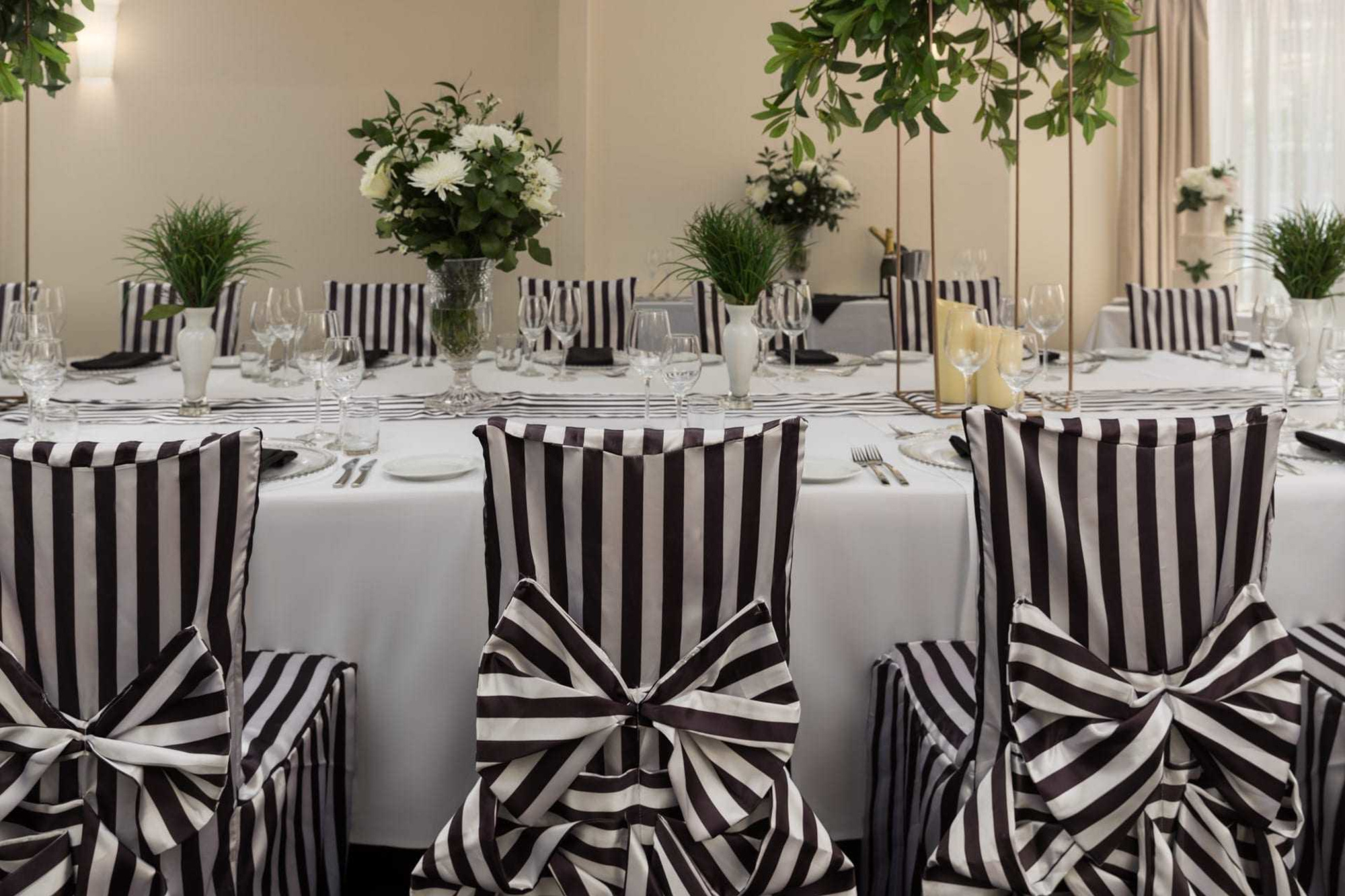 Elegant indoor wedding reception table with black and white striped chairs, glassware, and greenery centerpieces.