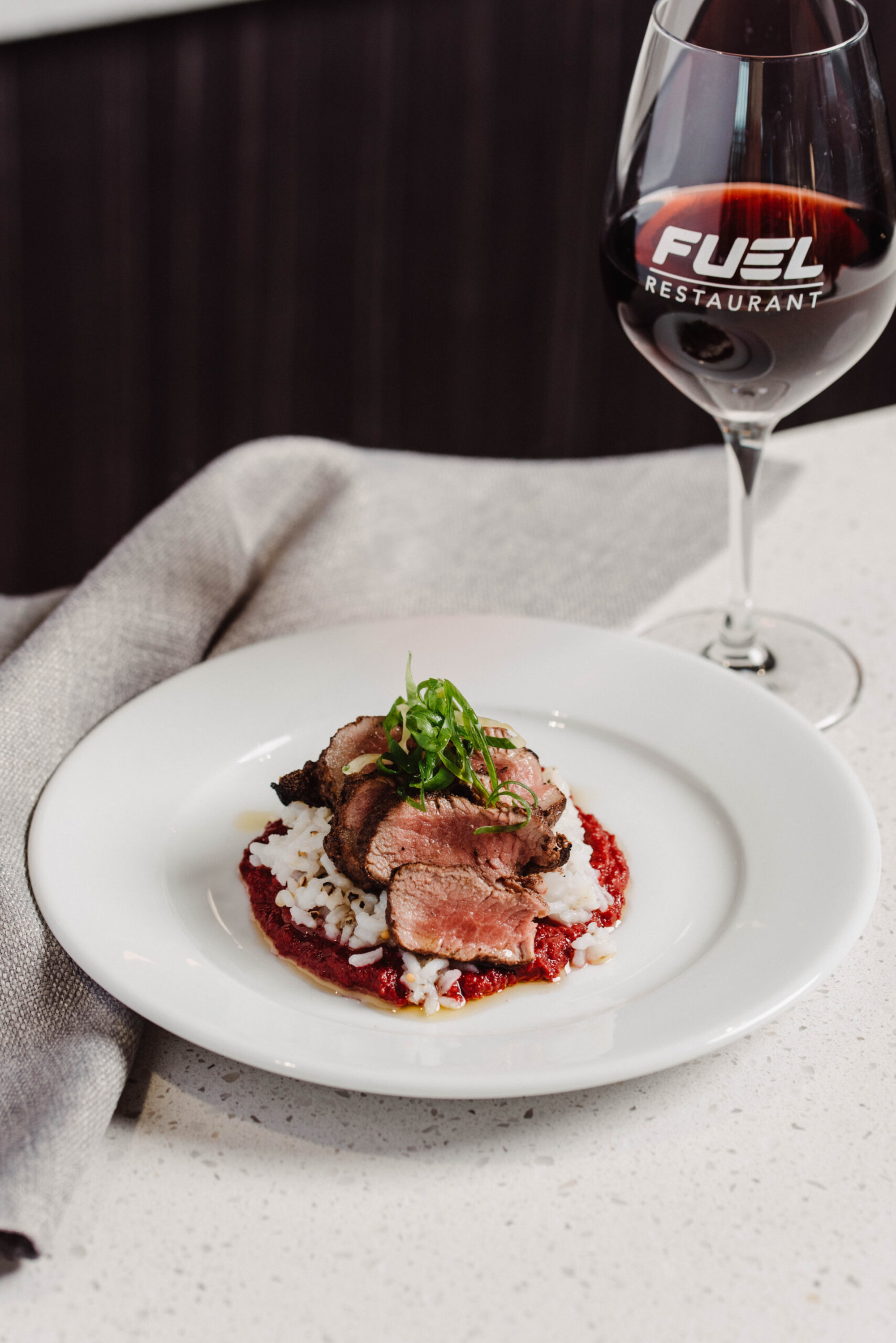 Gourmet plated steak over rice with sauce and a glass of red wine on a white table.