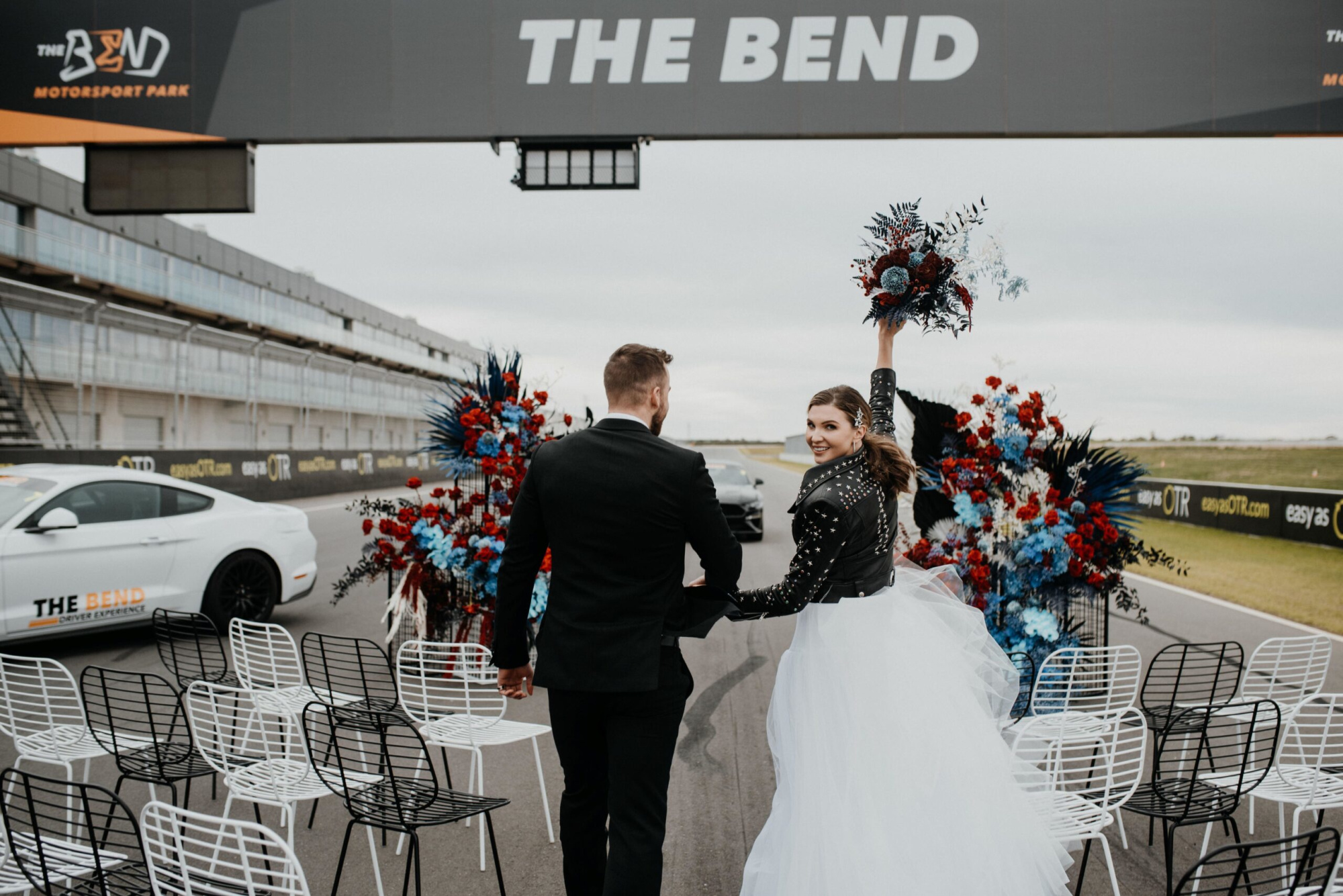 Bride and groom walk hand in hand down a racetrack aisle with bold red and blue florals at The Bend Motorsports Park.