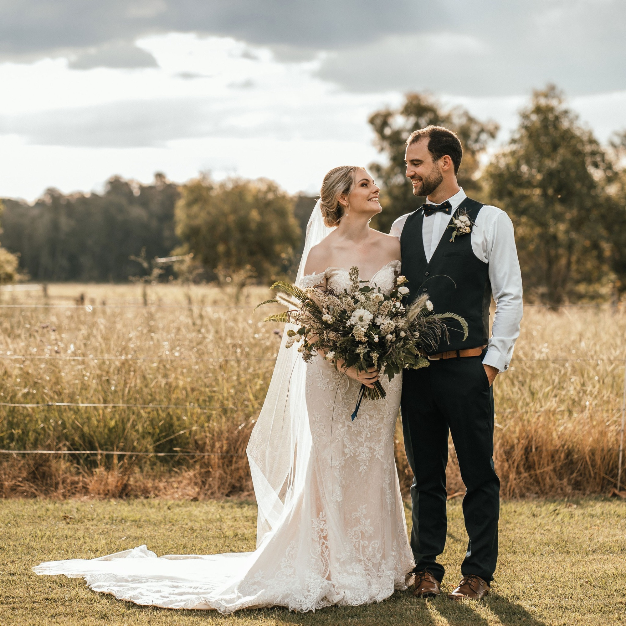 Bride and groom smiling at each other in a sunny field with a large rustic bouquet.