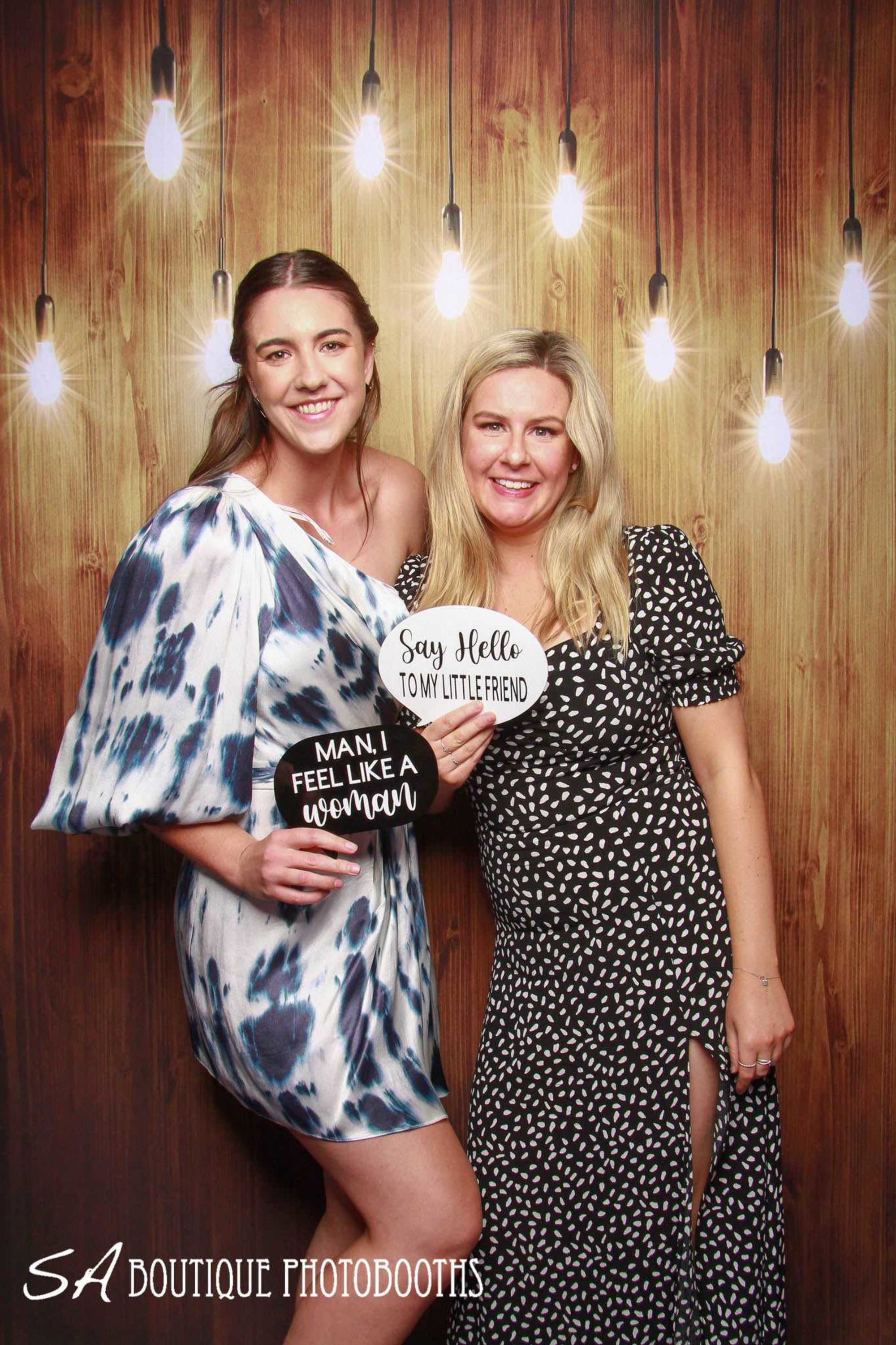 Two women pose with playful quote signs in front of a rustic wood and hanging bulb photo booth backdrop.