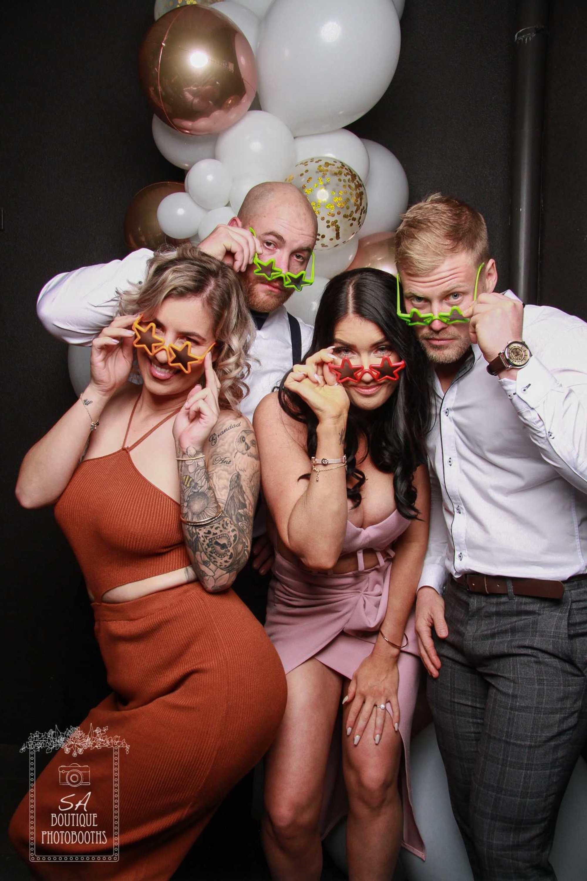 Four wedding guests pose playfully with star-shaped glasses in front of a white and gold balloon backdrop.