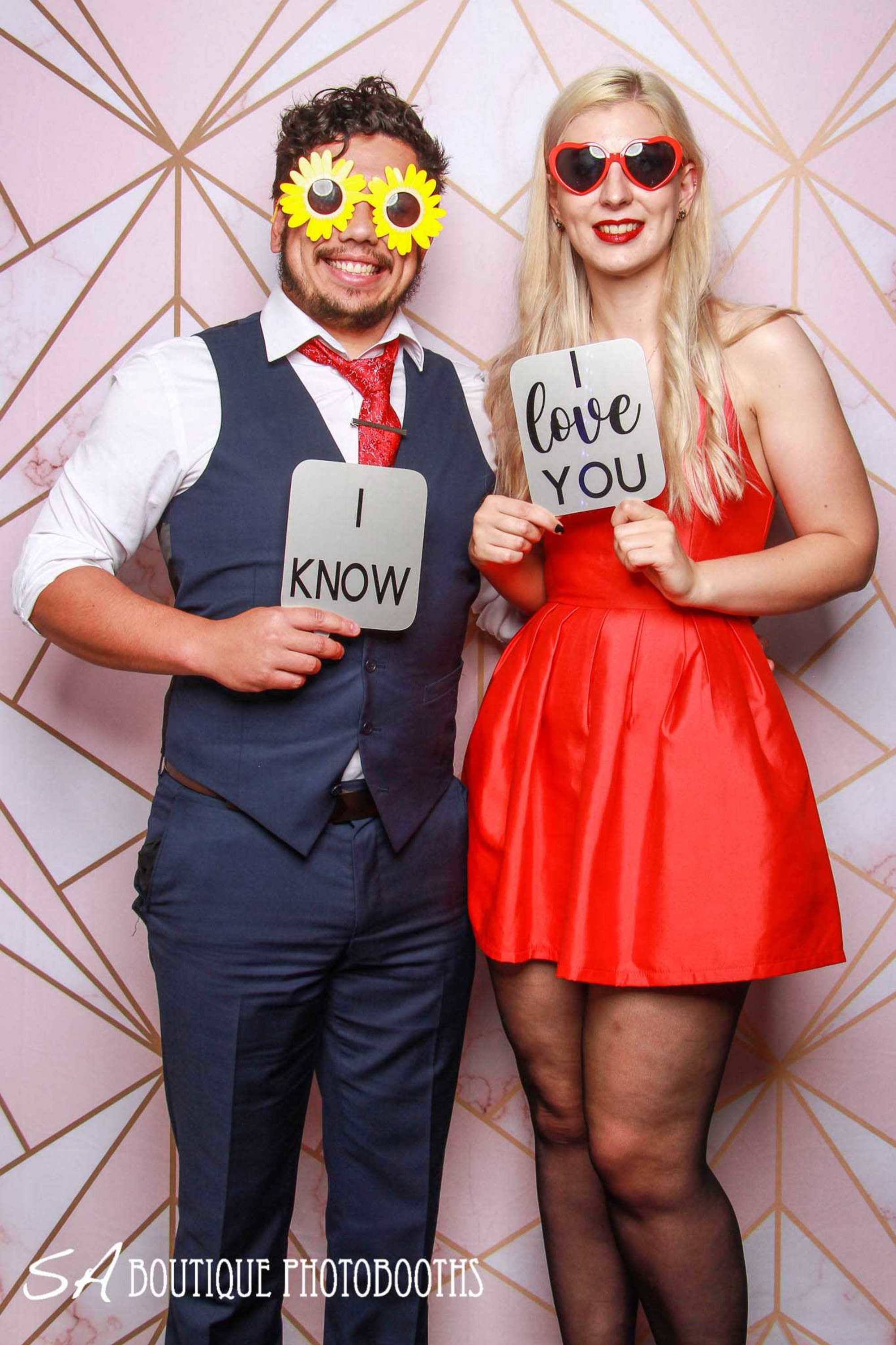 Smiling wedding guests pose in a photobooth with playful glasses and love-themed signs.