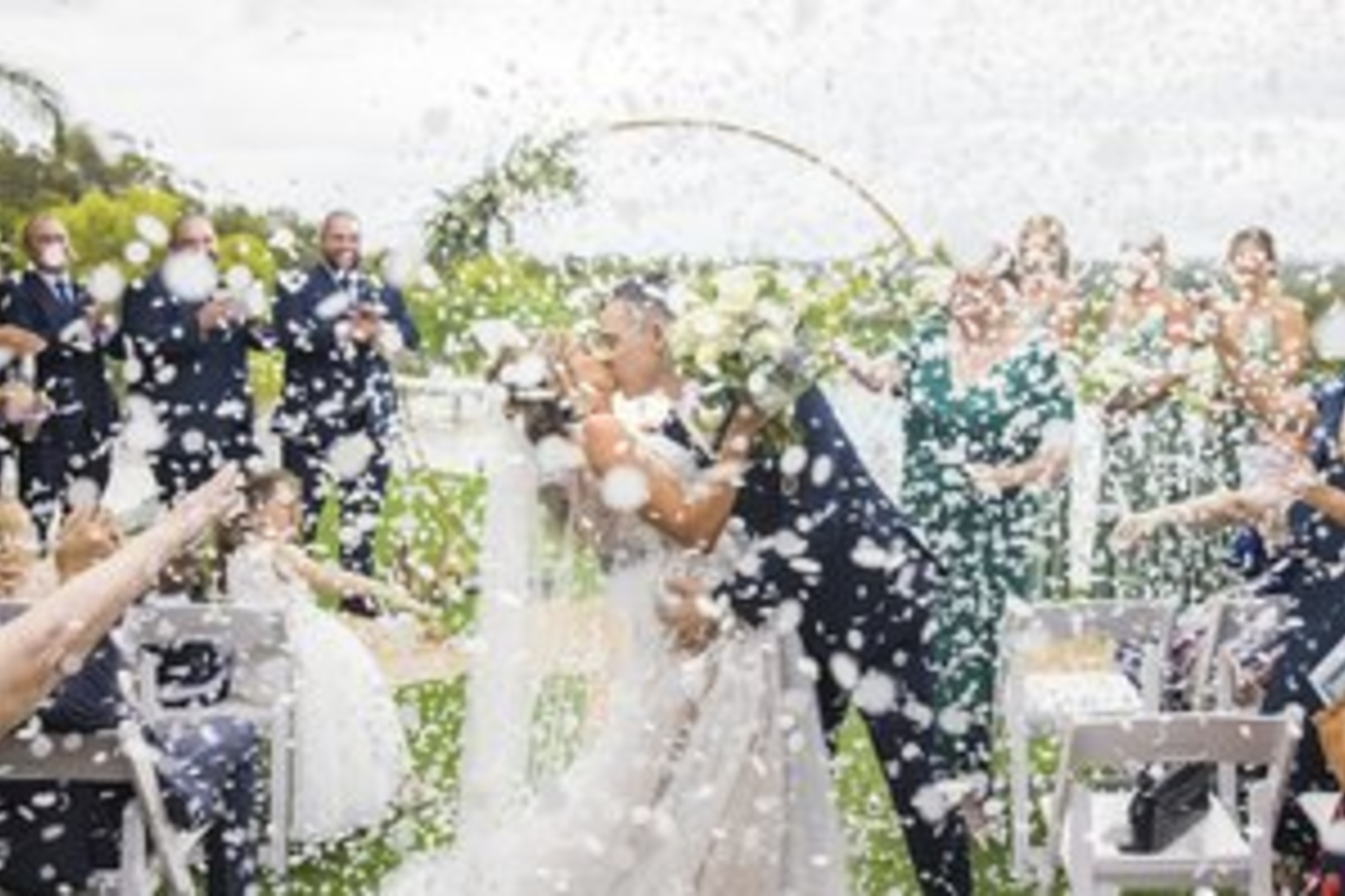 Newlyweds share a kiss during an outdoor ceremony exit as guests shower them with white confetti.