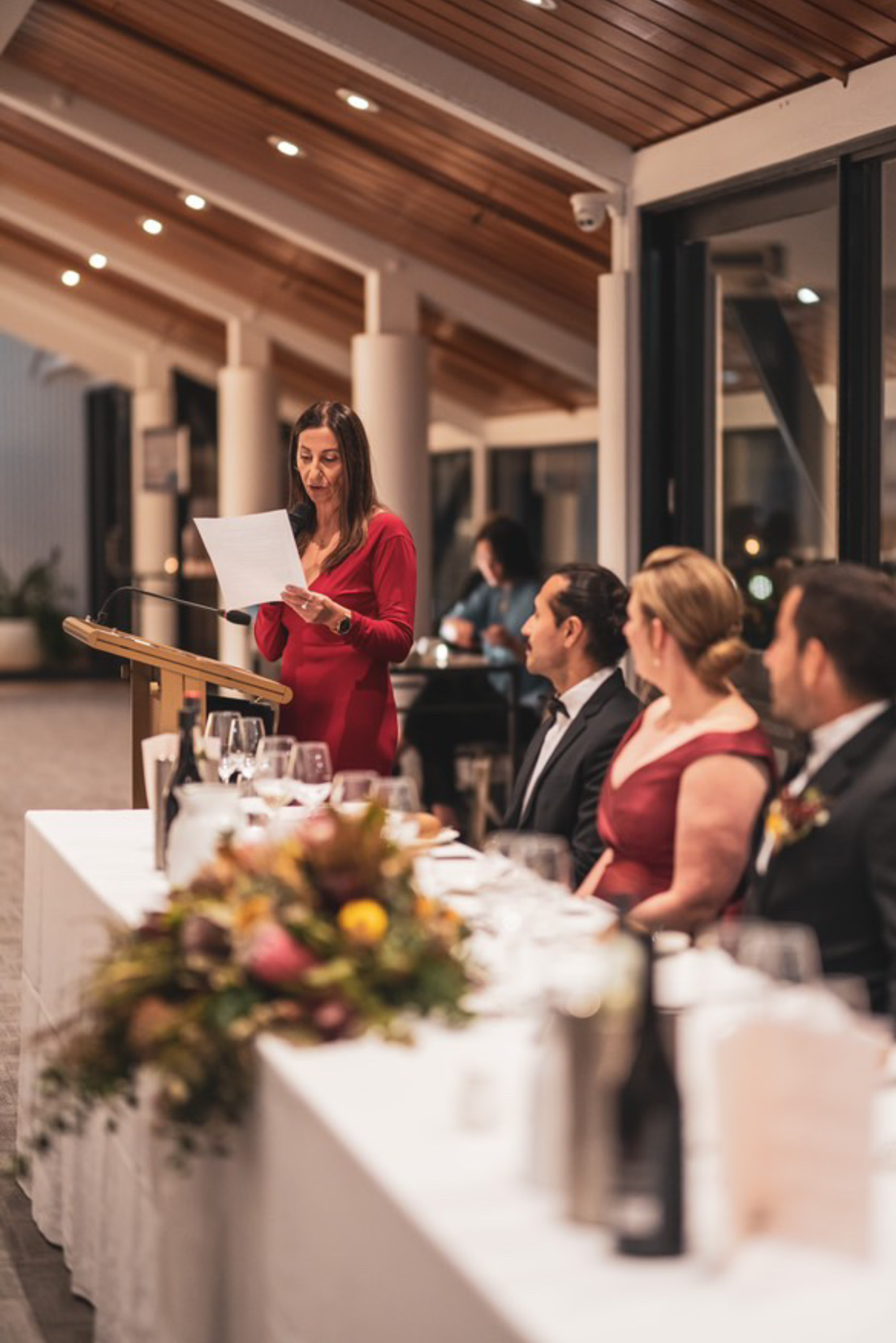 Woman in red dress gives a wedding speech at the reception head table in a modern indoor venue.