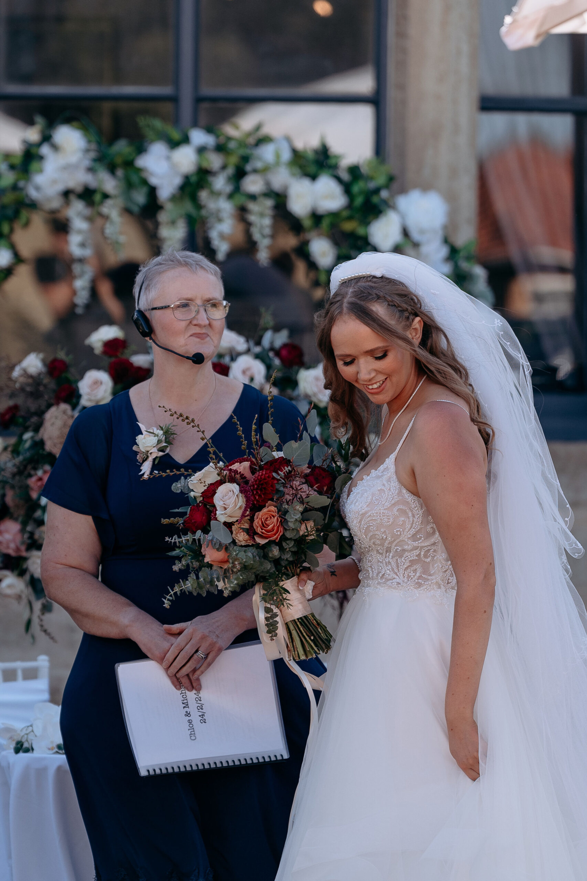 Bride holding a colorful bouquet stands beside the officiant during an outdoor wedding ceremony with floral backdrop.