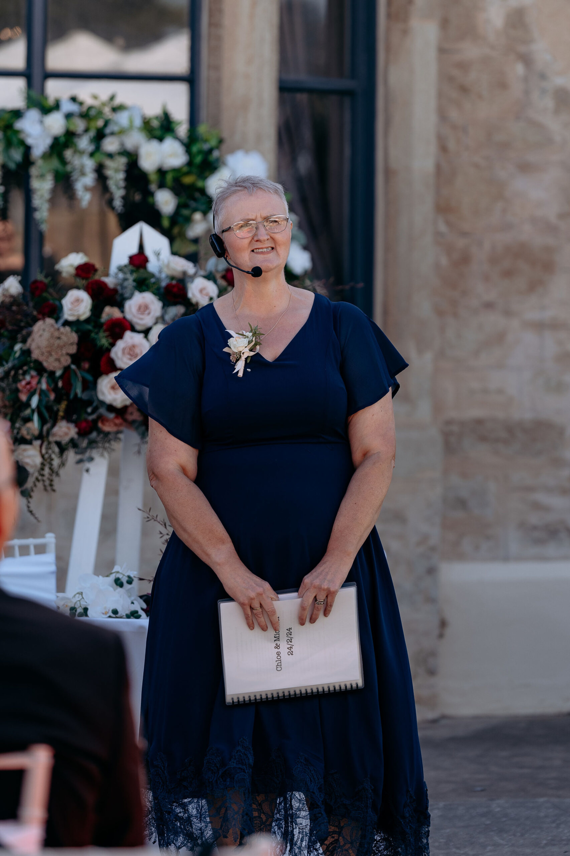 Wedding celebrant in a navy dress speaks into a headset microphone in front of a floral ceremony backdrop.