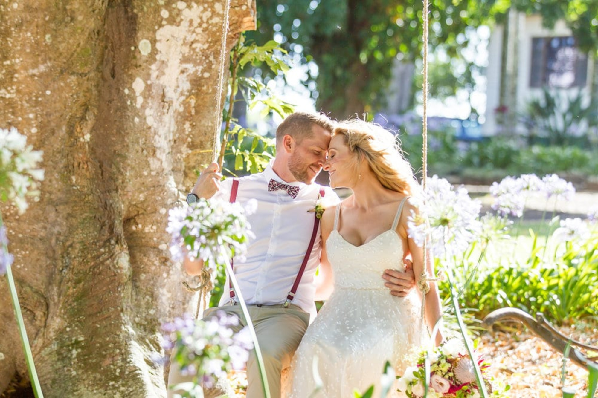 Bride and groom sit closely together on a flower-adorned swing in a sunny garden.
