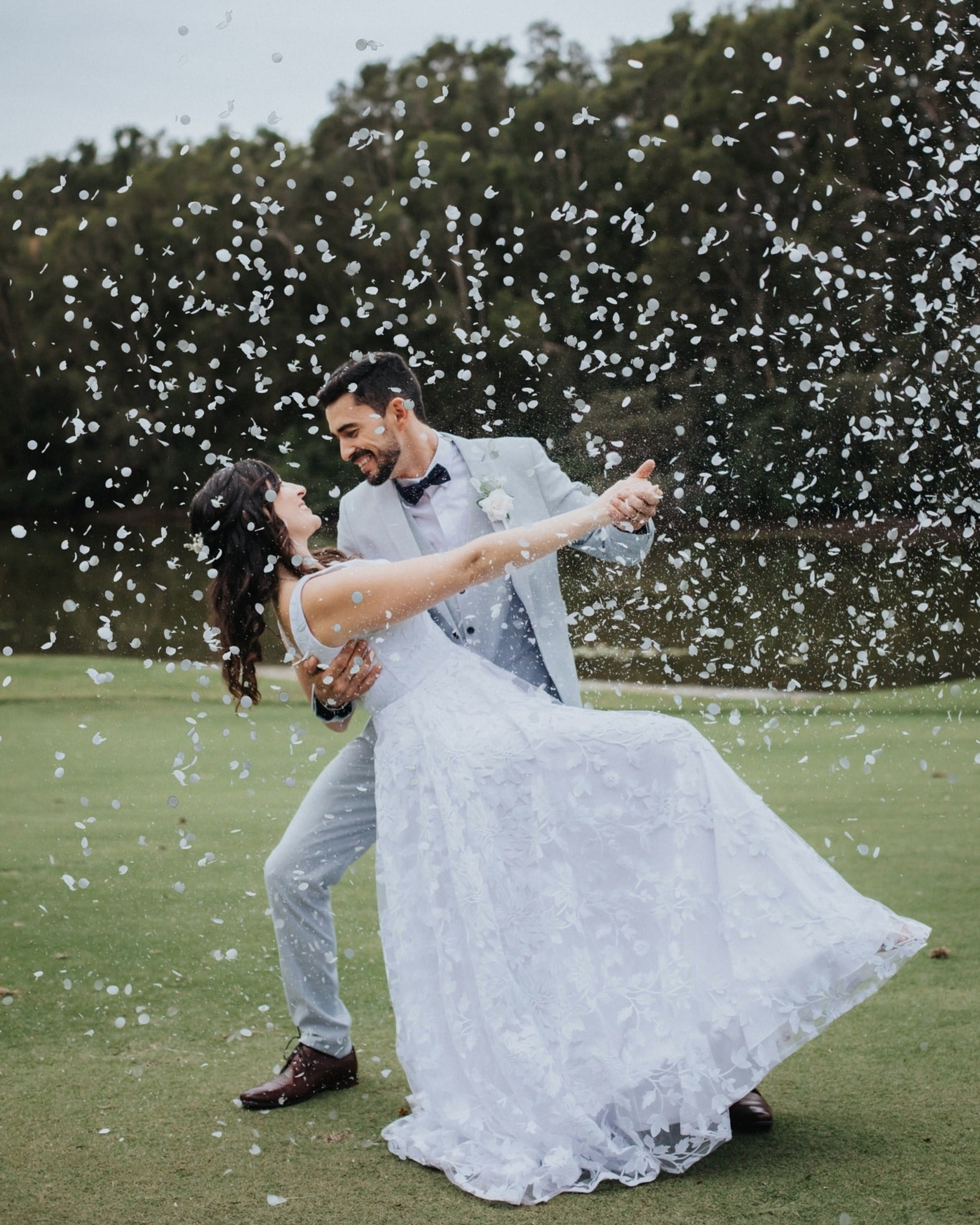 Newlywed couple dancing on a lawn as white confetti falls around them.