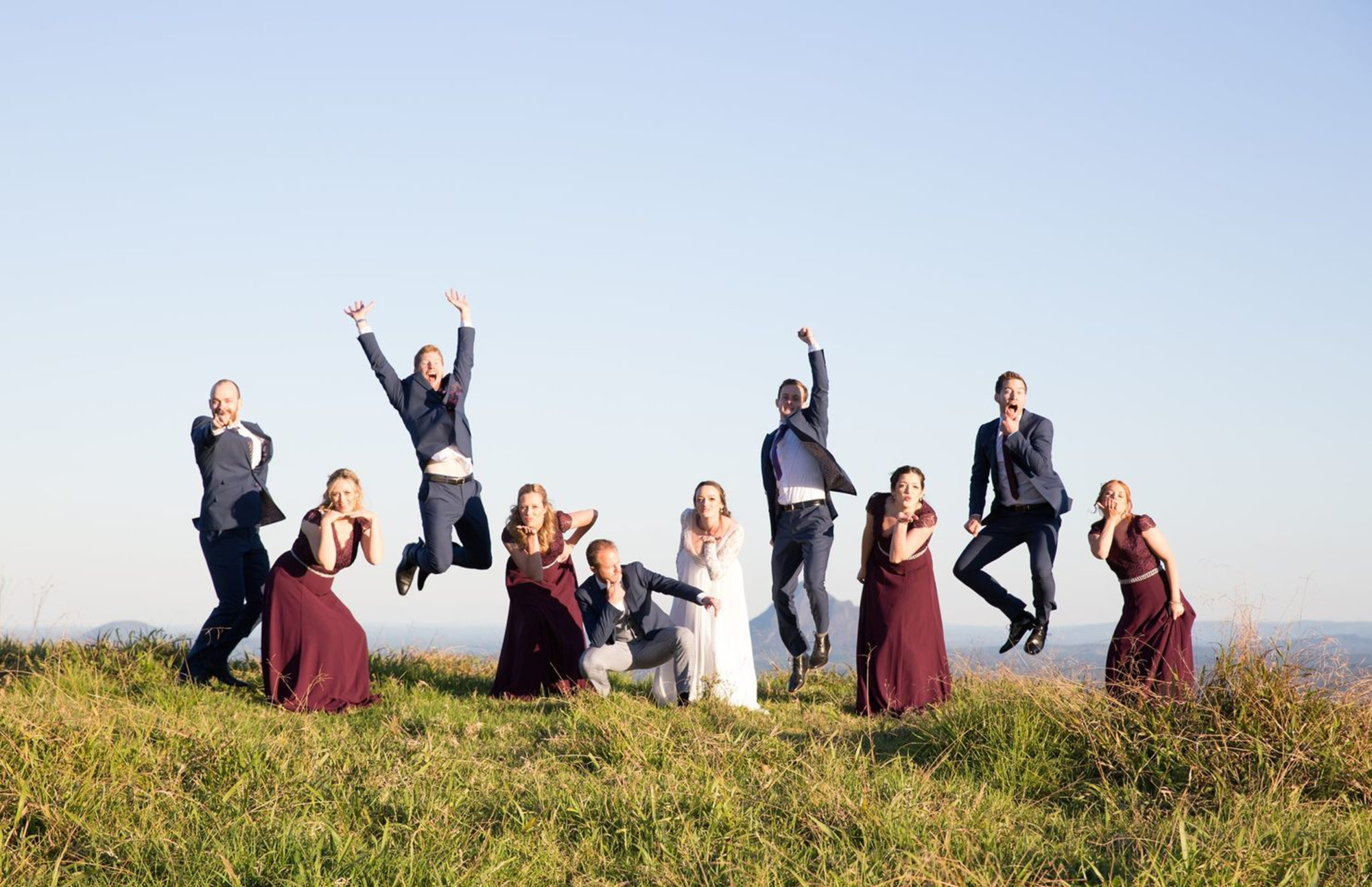 Joyful wedding party posing and jumping together on a grassy hill under a clear sky.