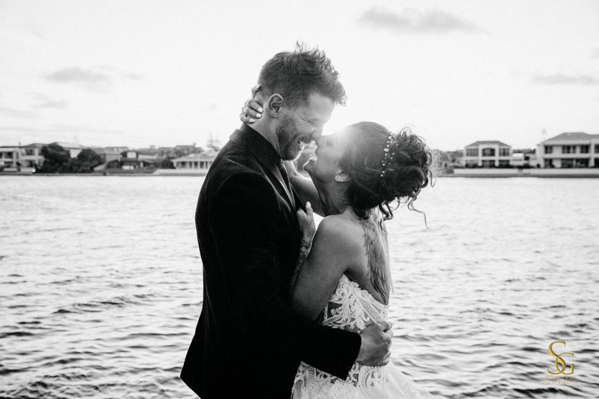 A newlywed couple embrace and laugh by the waterfront in a candid black and white wedding portrait.