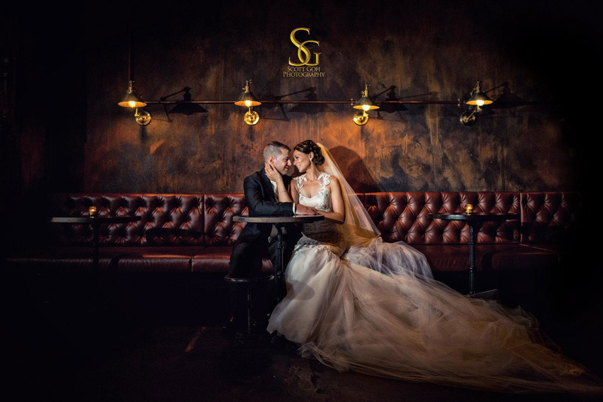 Bride and groom share an intimate moment on a tufted leather sofa in a dimly lit, moody lounge.