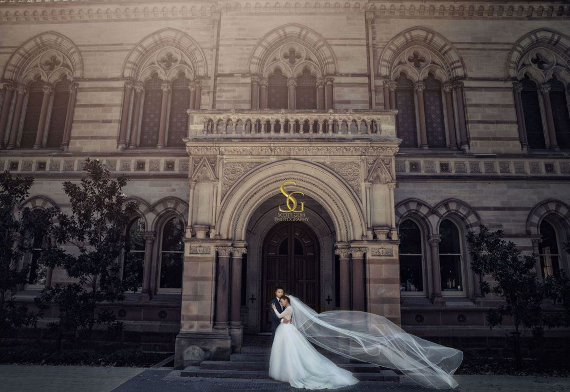 Bride and groom embrace in front of a grand historic stone building with the bride’s veil flowing in the wind.