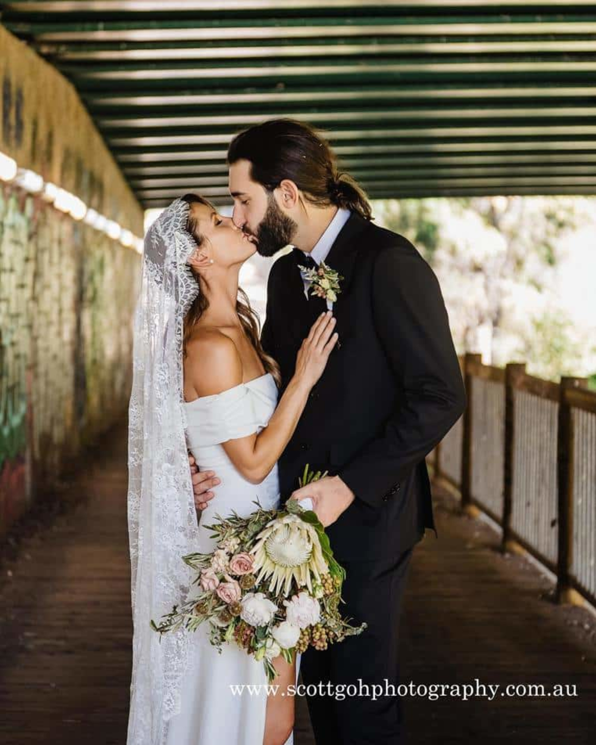 Bride and groom share a kiss in a tunnel while holding a natural bouquet.