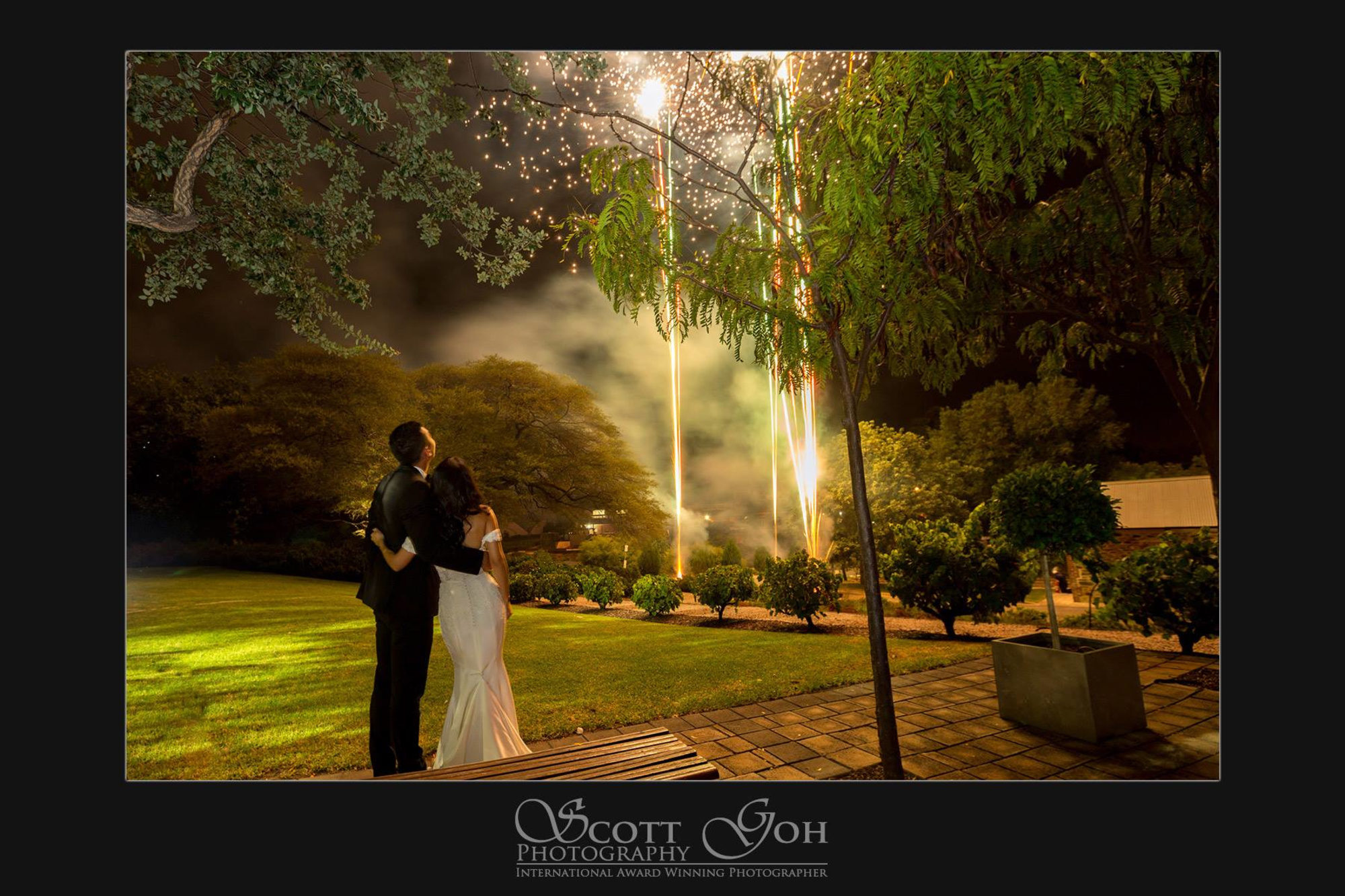 Bride and groom embrace in a garden at night watching fireworks burst in the sky.