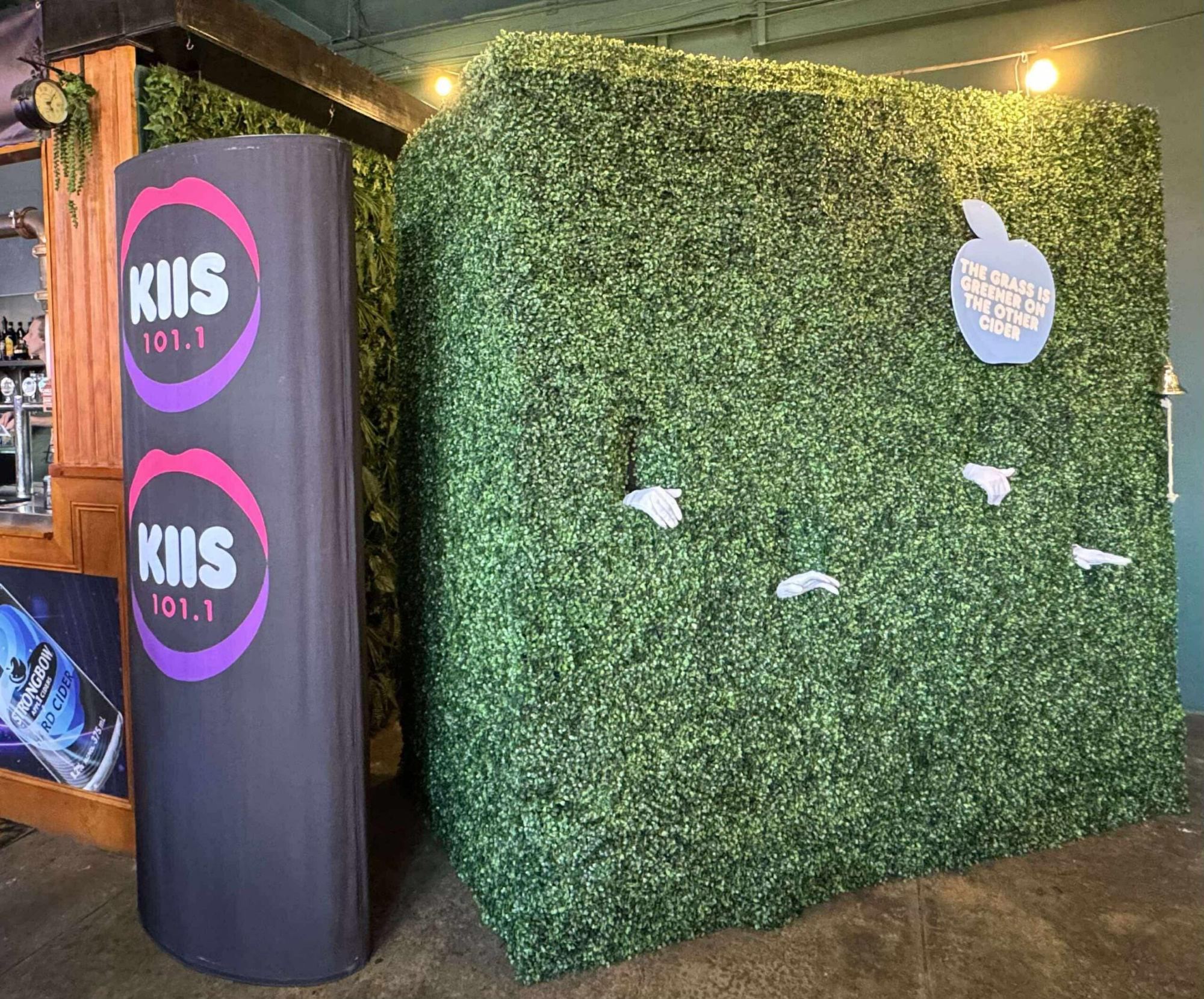 Greenery hedge photo booth with branded column and white glove props in an indoor venue.