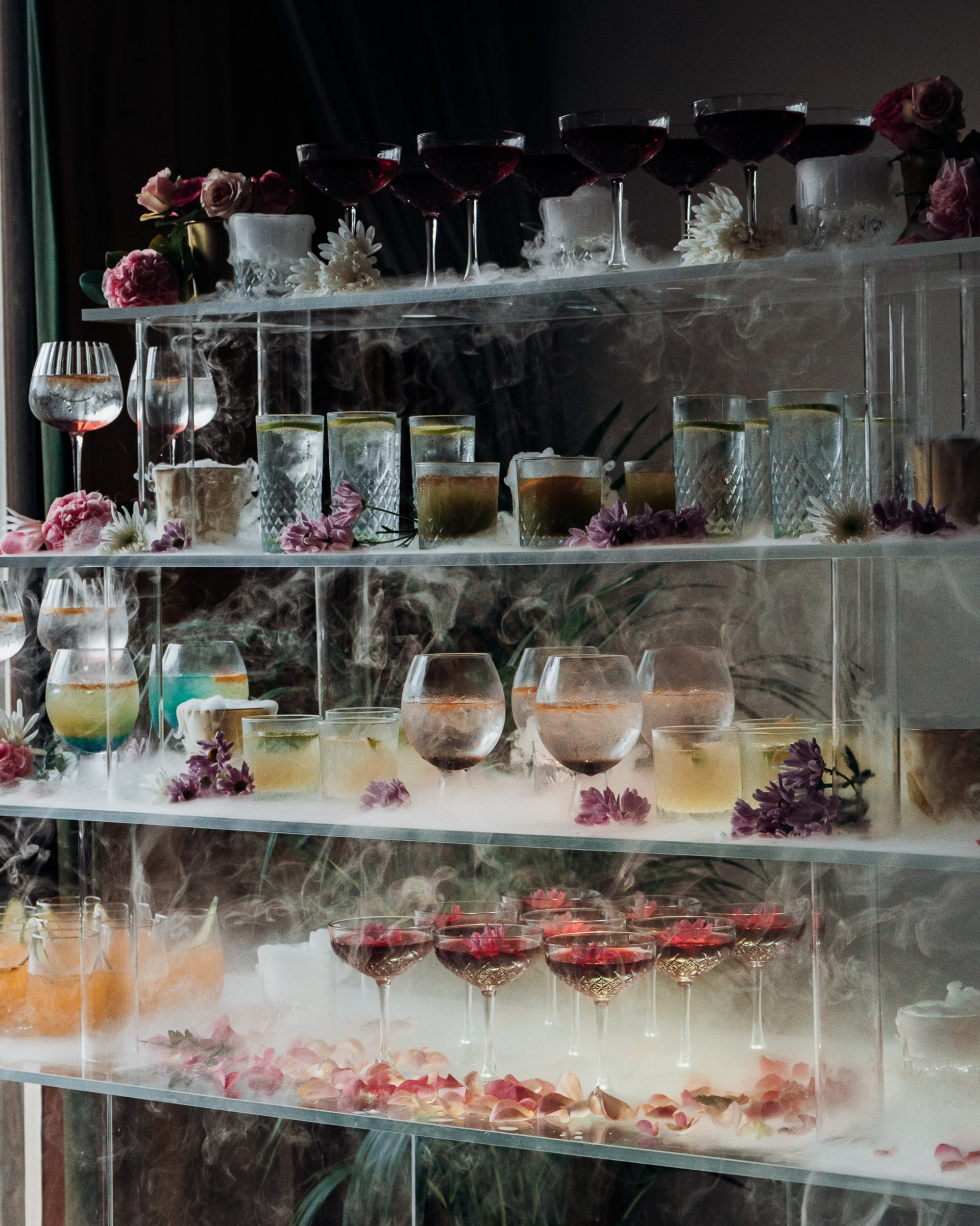 Elegant multi-tiered wedding cocktail display with smoky dry ice, flowers, and colorful drinks in glassware.