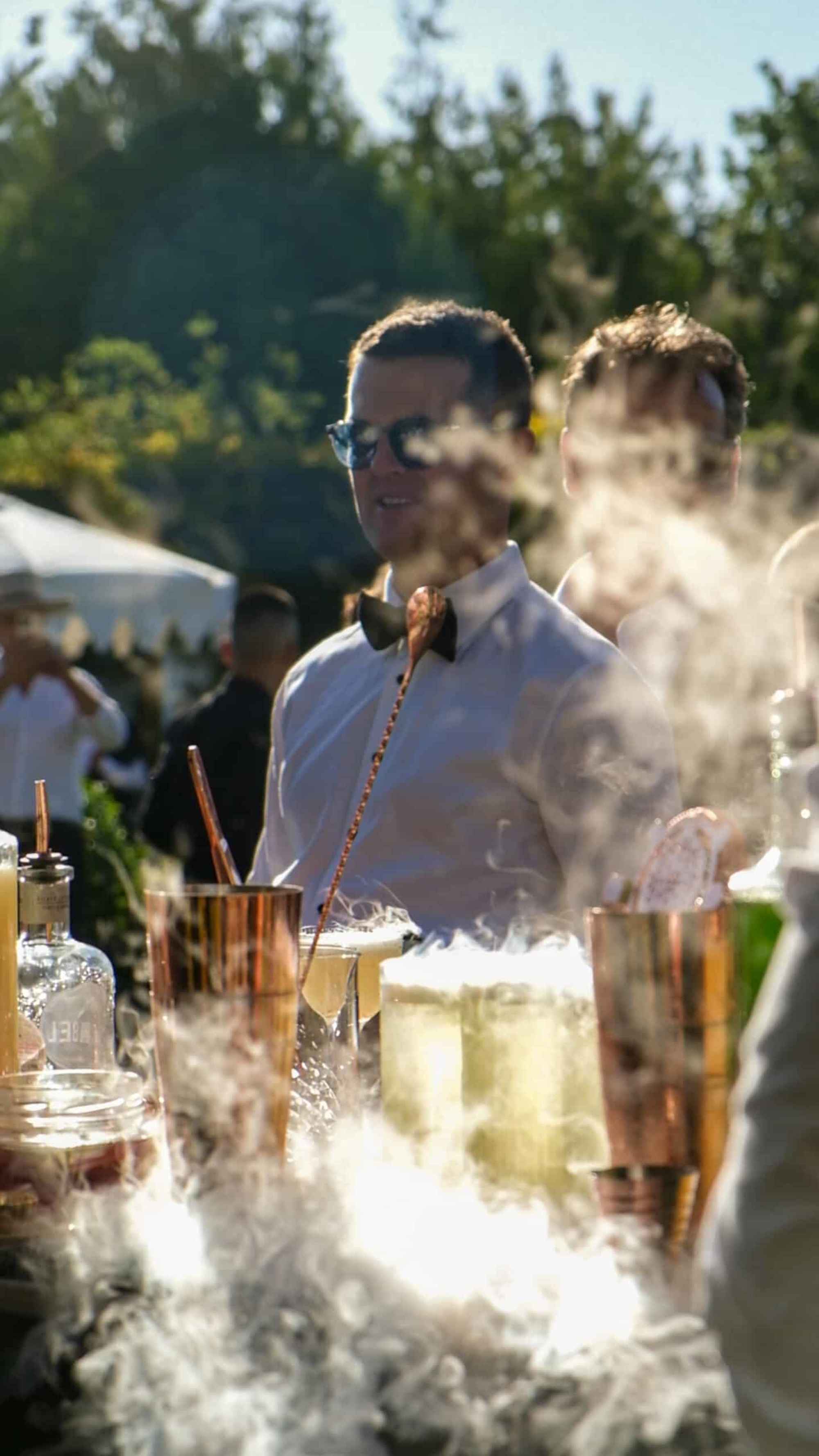 Bartender serving smoky signature cocktails at an outdoor wedding reception bar.