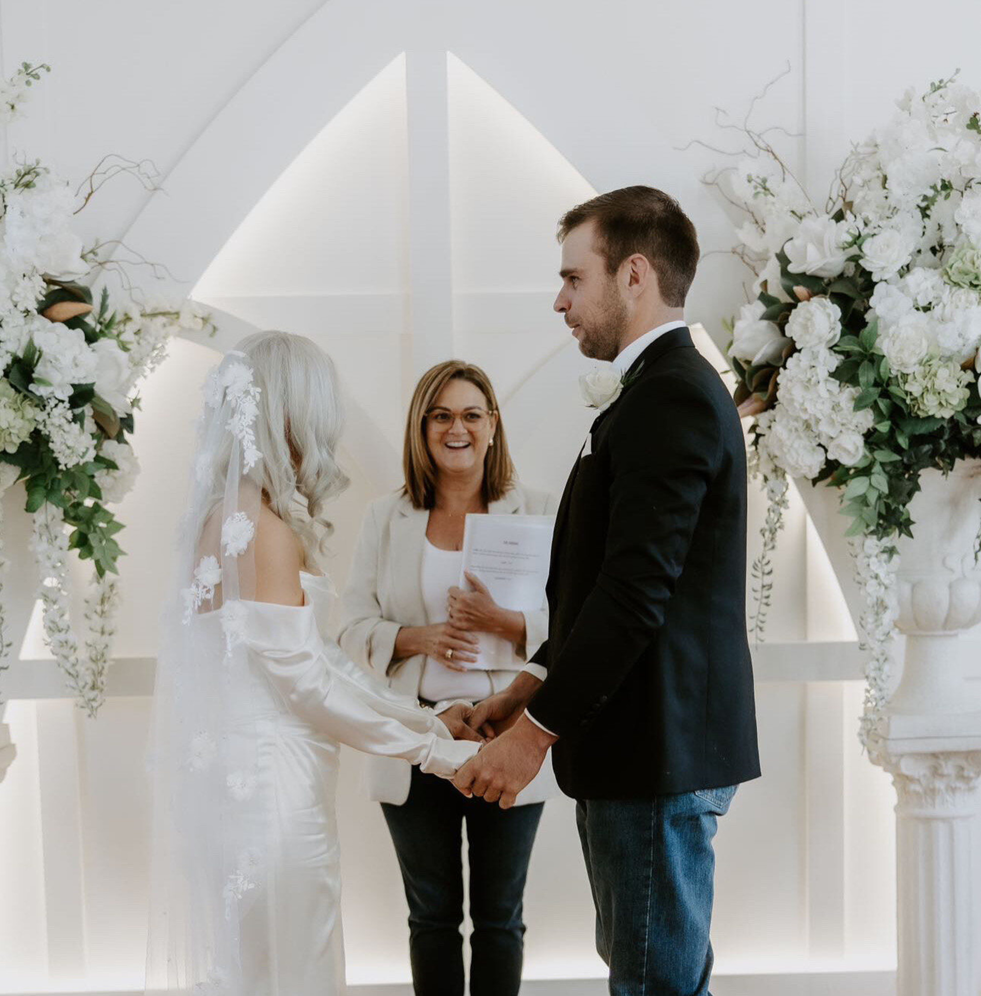 Couple holding hands during an indoor wedding ceremony with an officiant and white floral arrangements.
