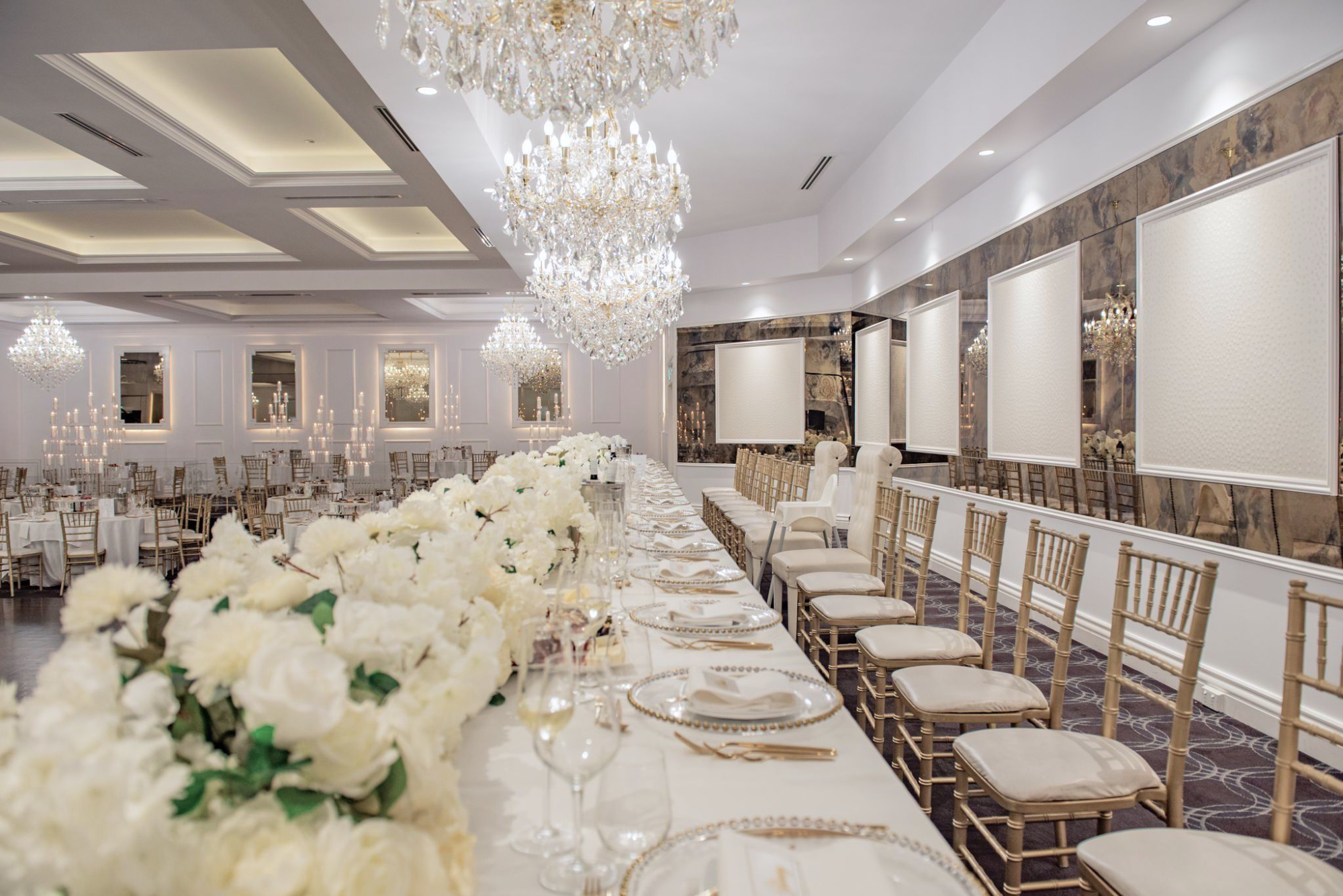 Elegant indoor wedding reception hall with long floral banquet table, gold chairs, and sparkling crystal chandeliers.
