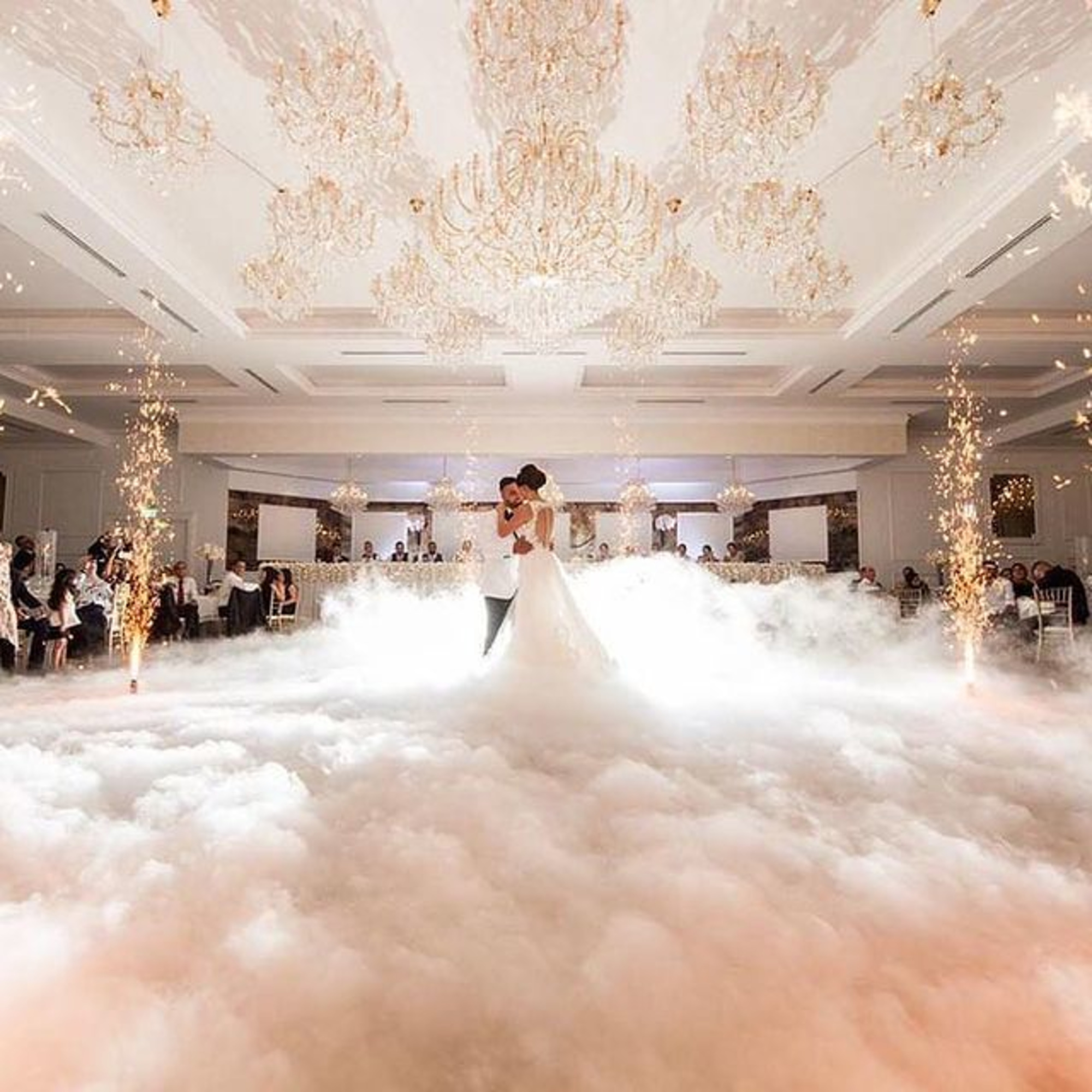 Bride and groom share a first dance in a fog-filled ballroom with sparkling fountains and crystal chandeliers.
