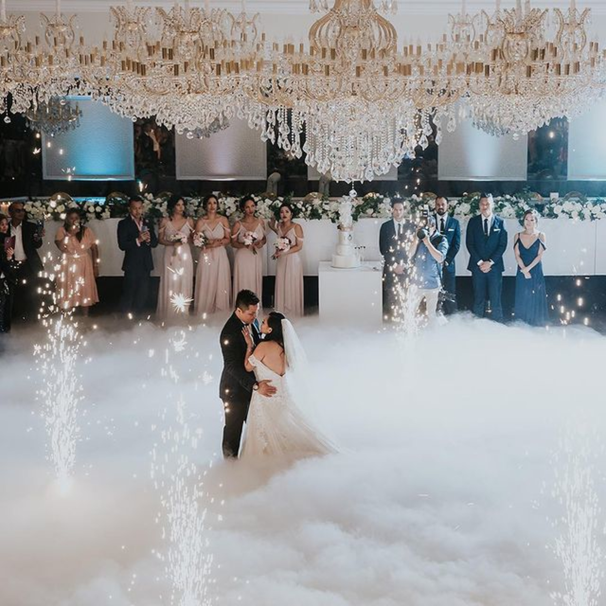 Bride and groom share a first dance amid fog and sparklers beneath grand chandeliers with their wedding party behind them.