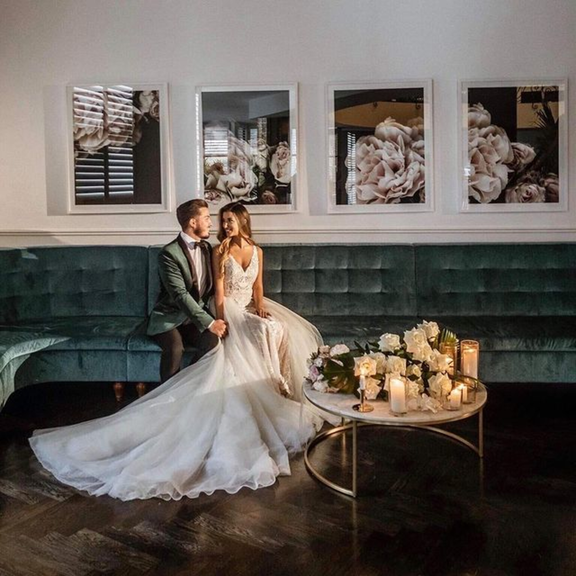 Bride and groom sit on a teal velvet sofa in a candlelit lounge with floral artwork and a flowing wedding dress train.