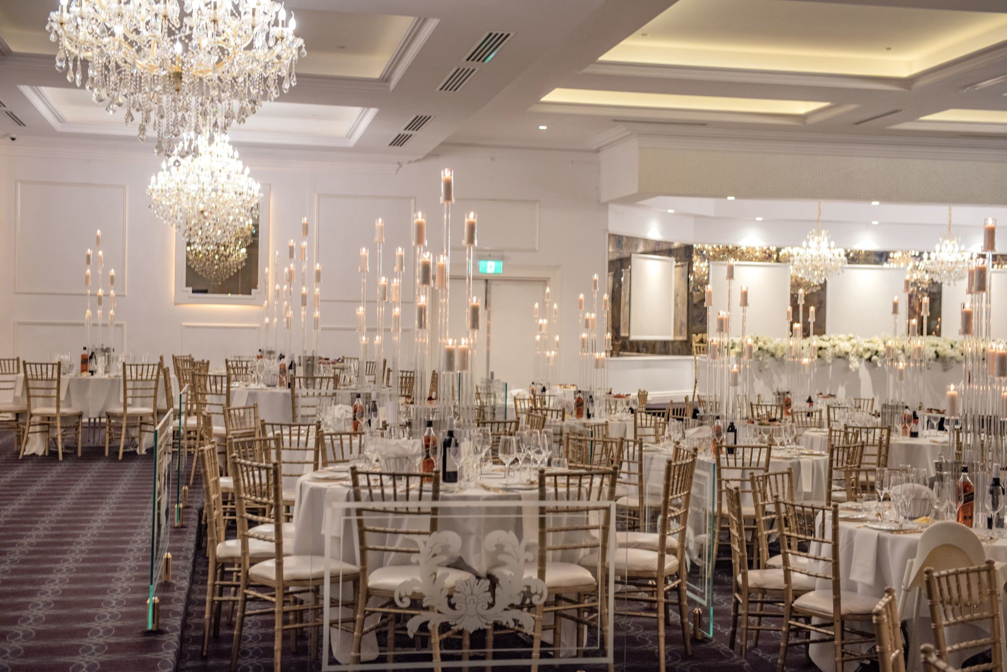 Elegant wedding reception ballroom with crystal chandeliers, tall candle centerpieces, and round tables with gold chairs.