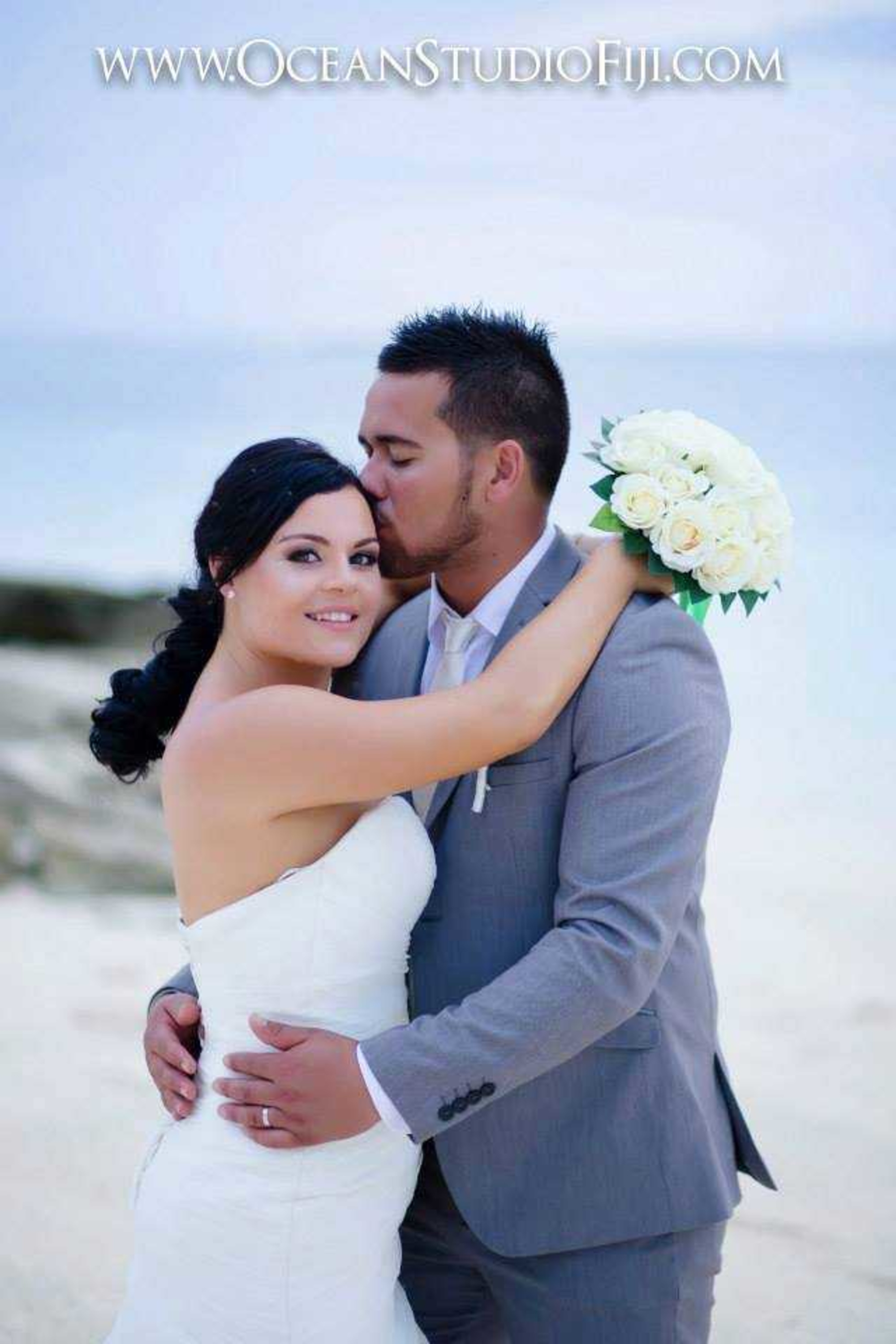 Bride and groom embrace on a sandy beach as he kisses her forehead, holding a white rose bouquet by the ocean.