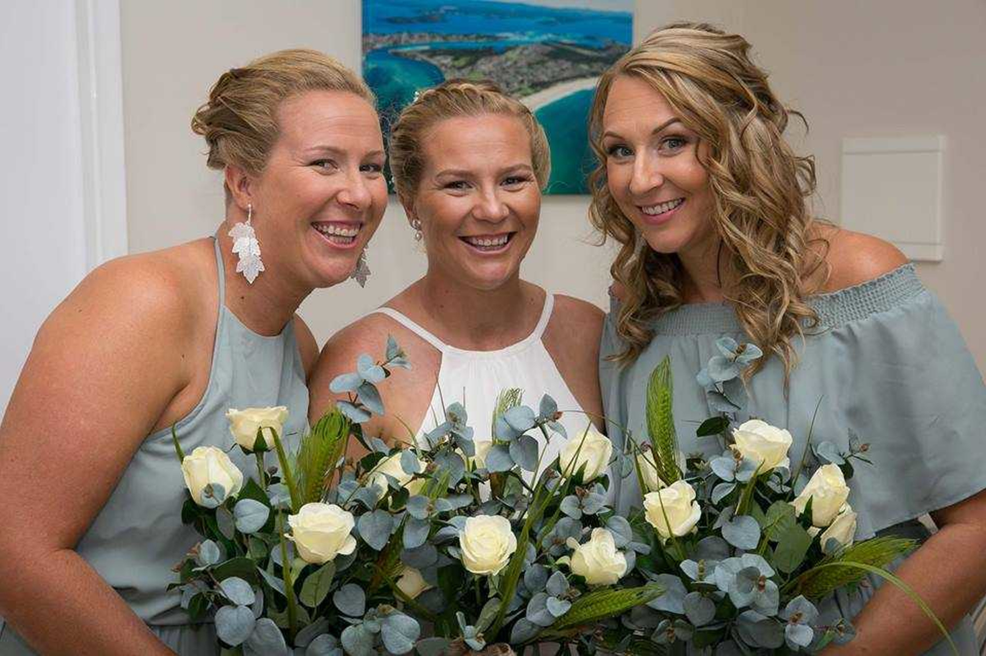 Bride and bridesmaids smiling indoors while holding pastel green and white wedding bouquets.
