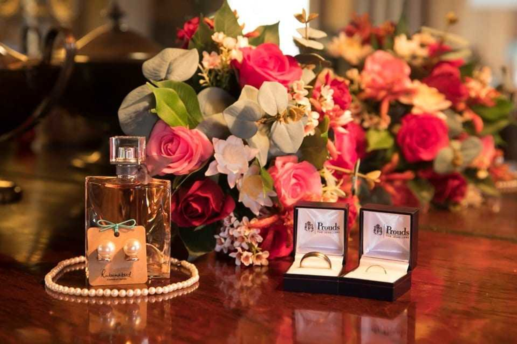 Wedding rings, perfume, pearls, and a romantic rose bouquet arranged on a polished wooden table.
