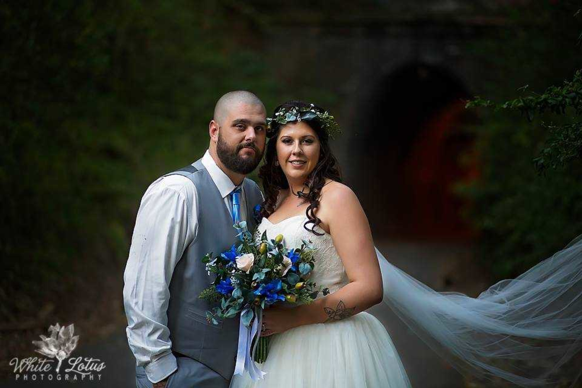 Bride and groom pose outdoors with a flowing veil and vibrant bouquet in a wooded setting.