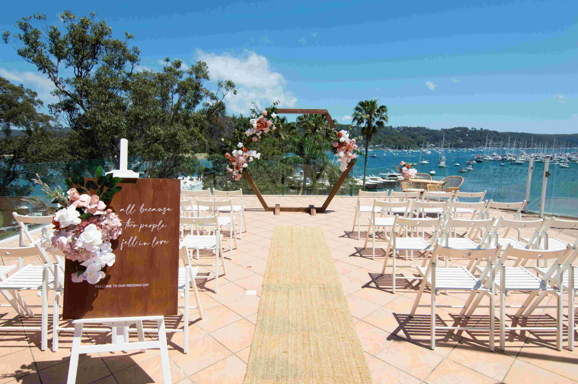 Outdoor waterfront wedding ceremony setup with floral hexagon arch, white chairs, and harbor views.