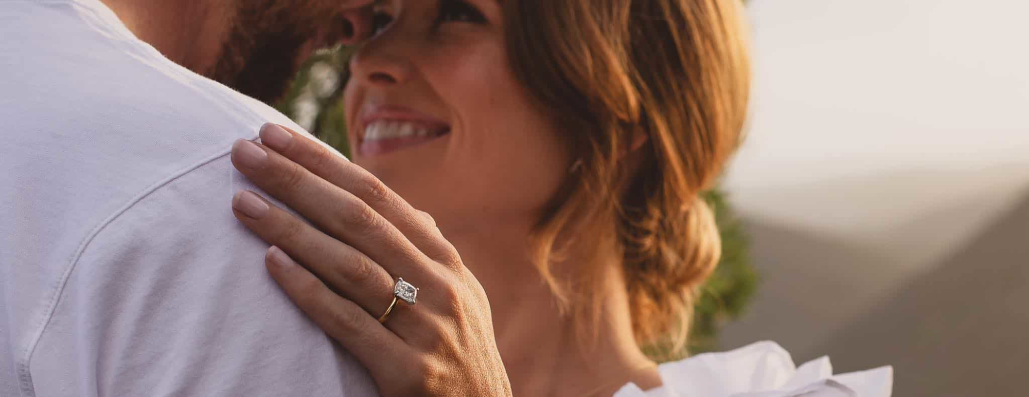 Close-up of a woman smiling and embracing her partner, showcasing a diamond engagement ring on her hand.