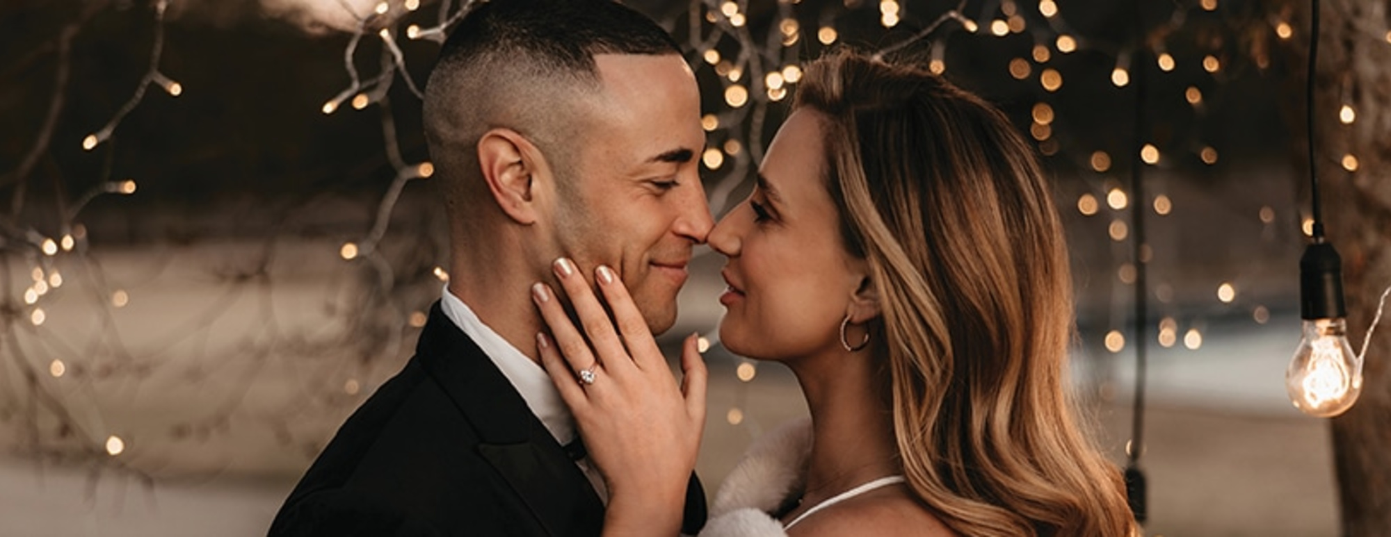 An intimate wedding couple portrait at dusk under warm string lights, highlighting the bride's engagement ring.