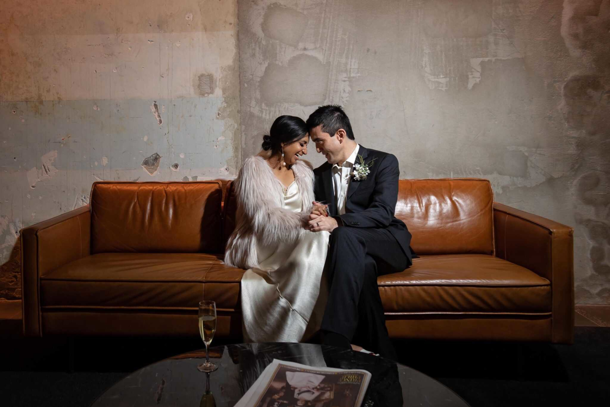 A newlywed couple sits closely on a leather sofa in an industrial-style room, holding hands and smiling.